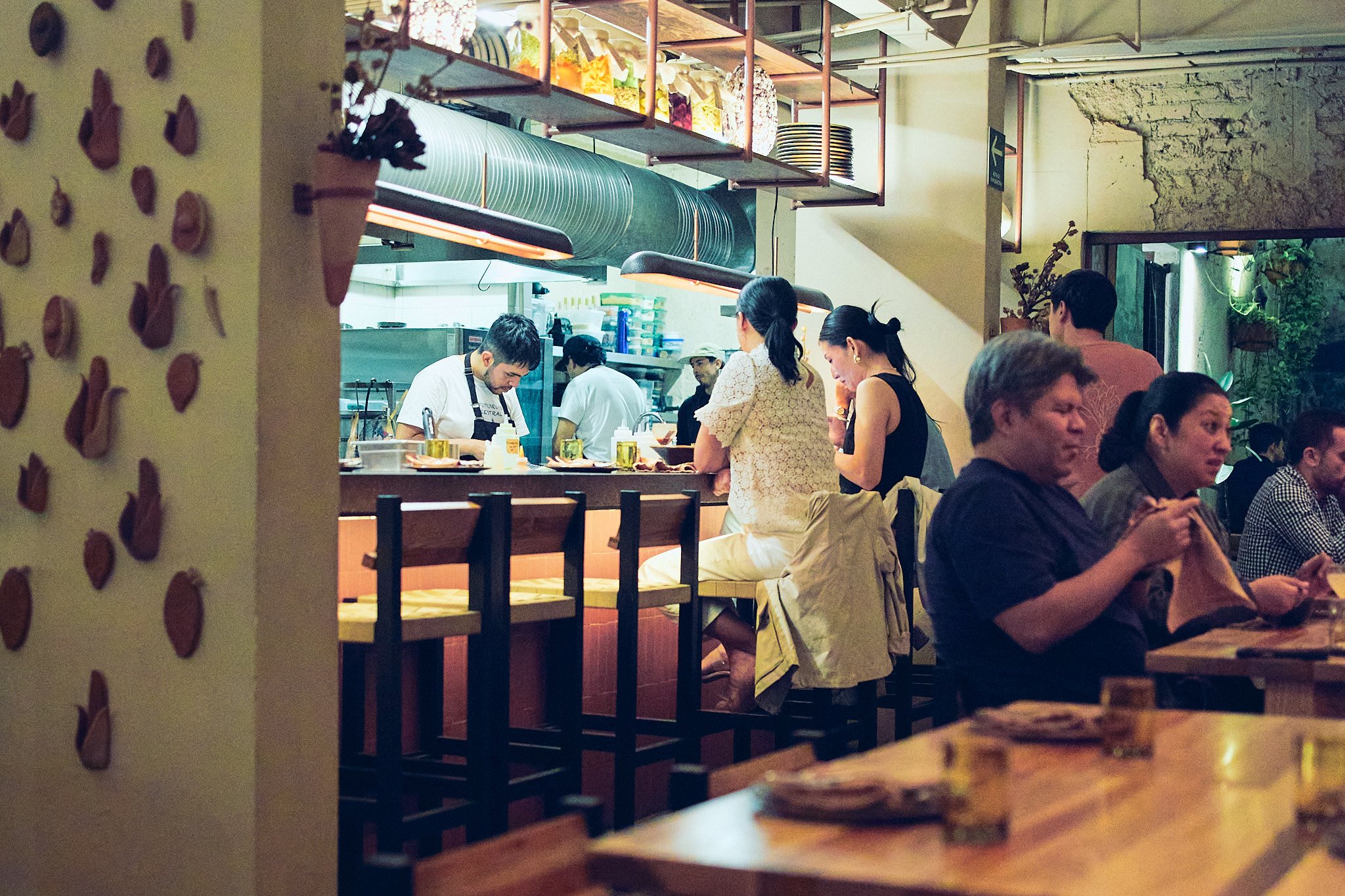 People sitting at tables inside a restaurant, with some chefs working behind a counter, and shelves with colorful containers and plates above.