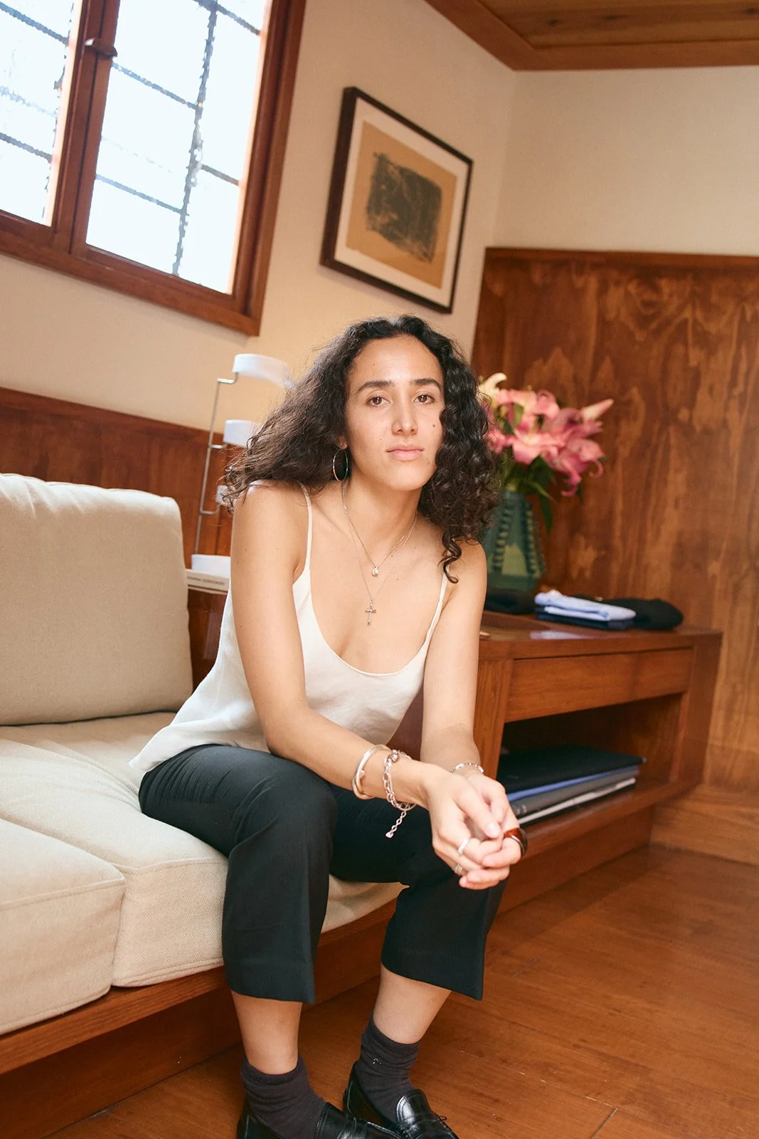 A young woman with curly dark hair sitting on a beige sofa in a room with wooden walls and a window, wearing a white sleeveless top, black pants, and black shoes, with jewelry including rings, bracelets, and necklaces, sitting with her hands clasped 
