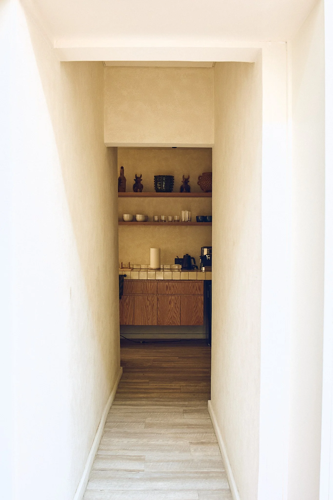 A narrow hallway leading to a small kitchen area with wood cabinets, a tiled countertop, cups, glasses, and kitchen appliances on the shelves.