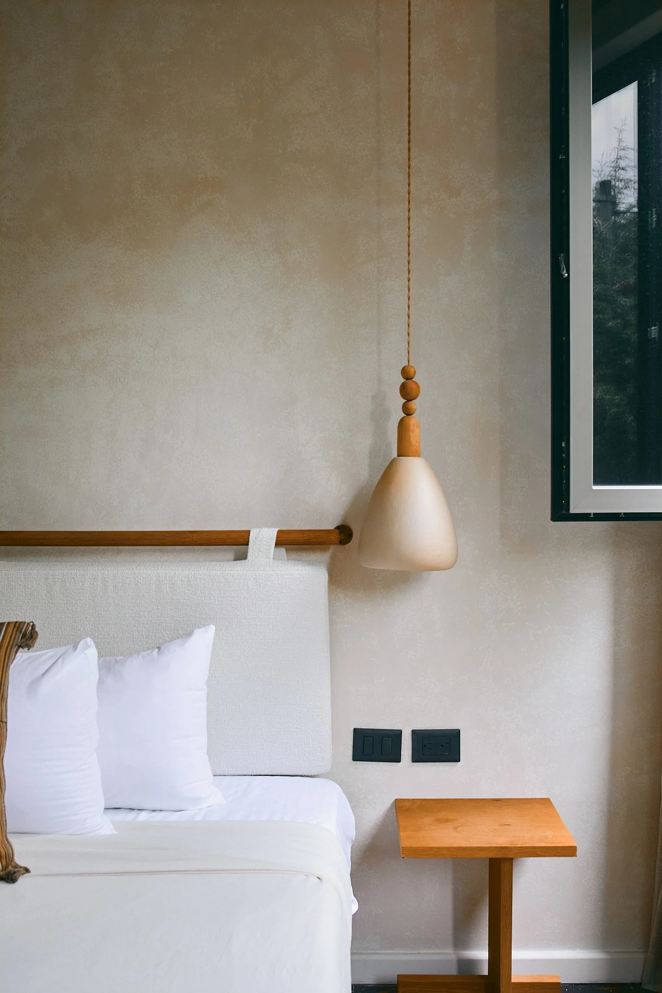 A bedroom corner featuring a bed with white pillows, a minimalist headboard, a beige wall art piece, and a hanging pendant light with a wooden detail.