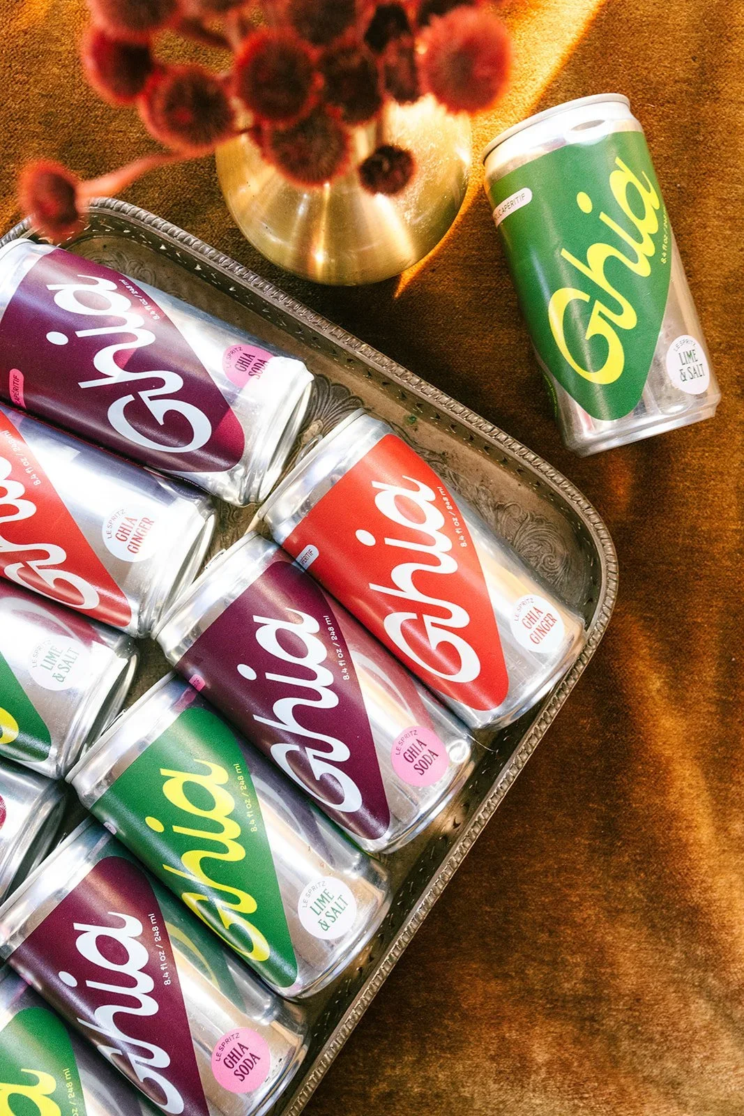 A silver tray filled with cans of Lime and Salt flavored soda, labeled 'Shw.' A single can with the same label is placed outside the tray on a brown surface, with a gold-colored vase of red flowers in the upper left corner.