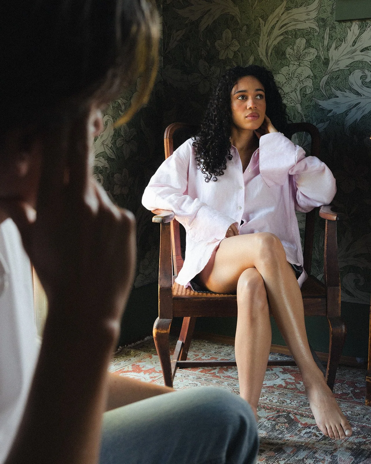 A woman with curly black hair sitting on a wooden chair, wearing an oversized pink shirt and black shorts, looking thoughtfully to the side in a room with floral wallpaper.