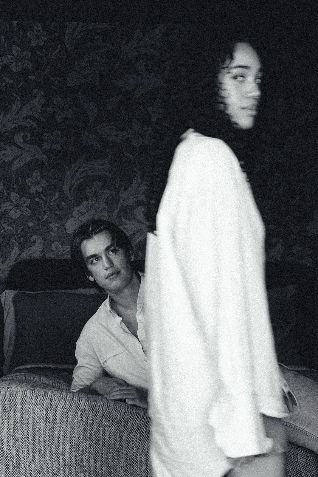 A black and white photo of a woman with curly hair standing in the foreground, looking to her right. In the background, a man is lying on a couch, looking at her, with a patterned wallpaper behind him.