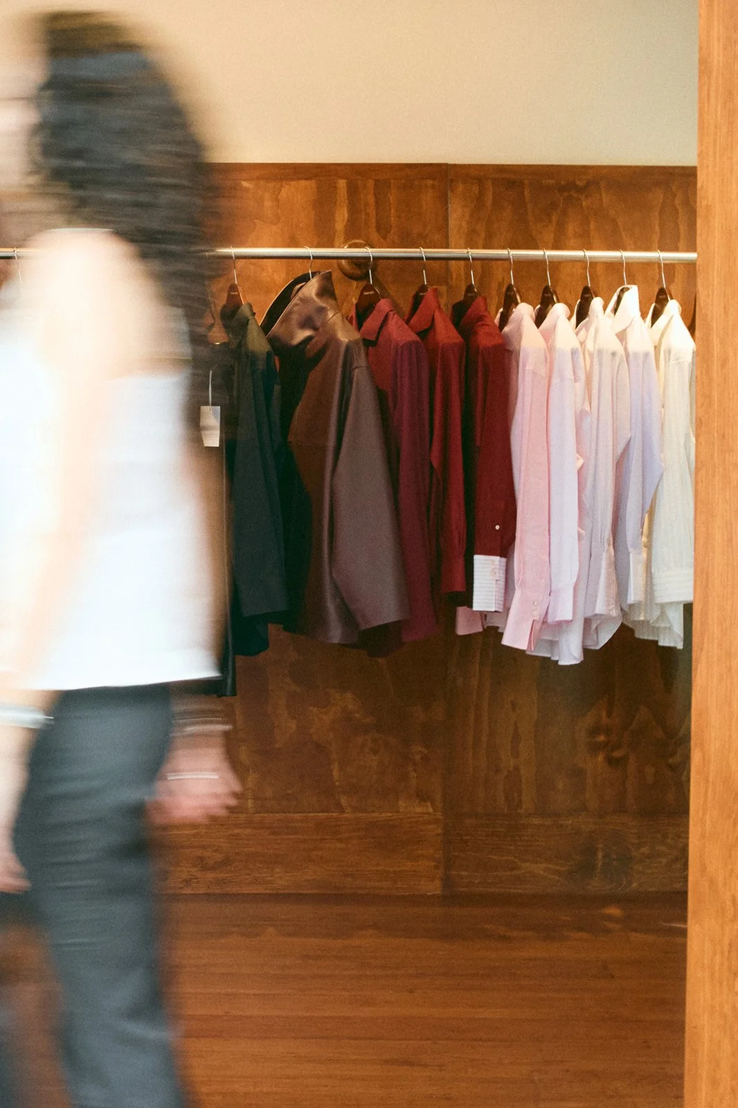 A person walking past a clothing rack with various shirts in black, brown, red, light pink, and white colors, in a store with wood-paneled walls.