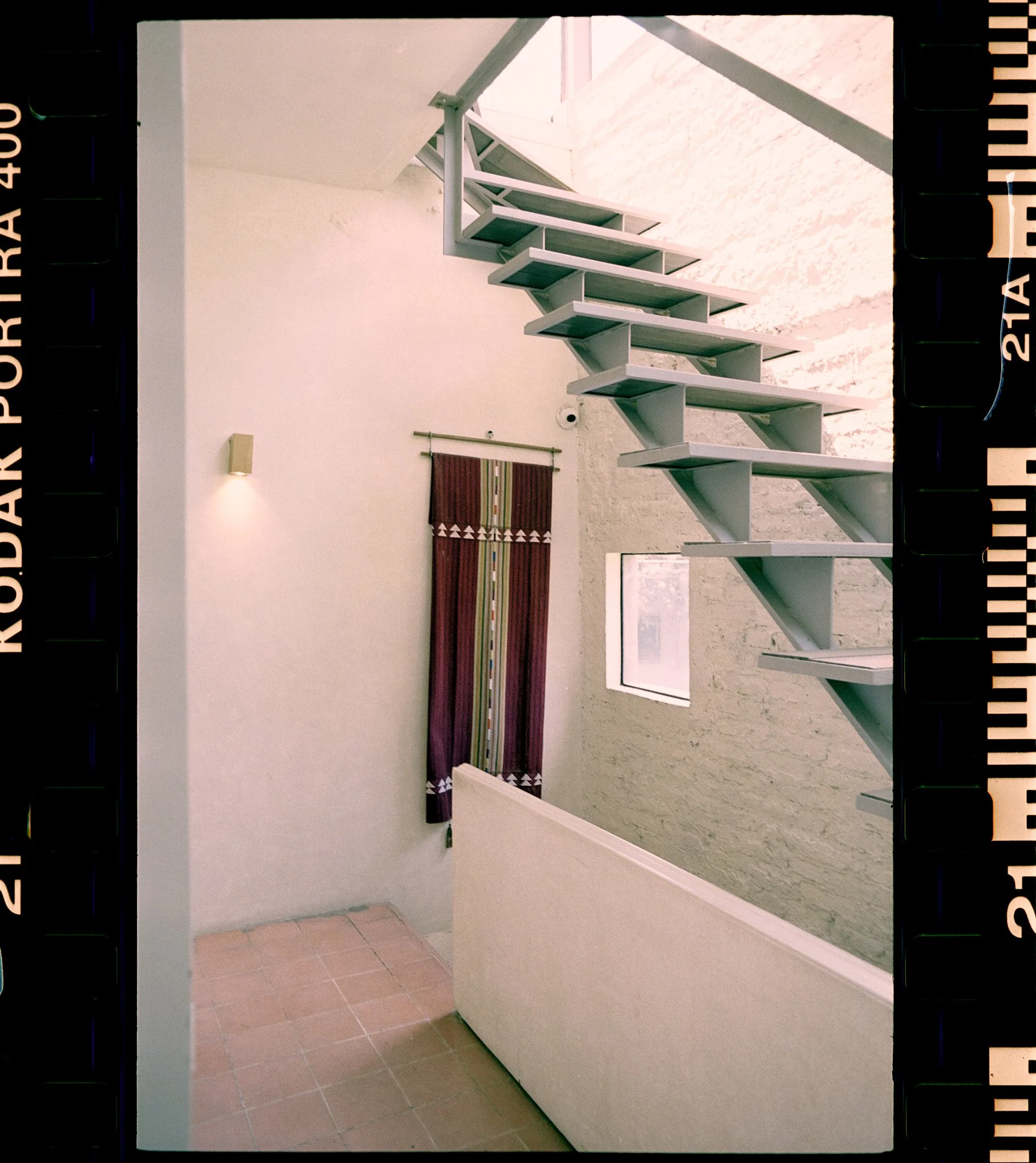 An indoor space featuring a modern metal staircase with open steps, a small window, a wall-mounted light fixture, and a patterned textile hanging on the wall.