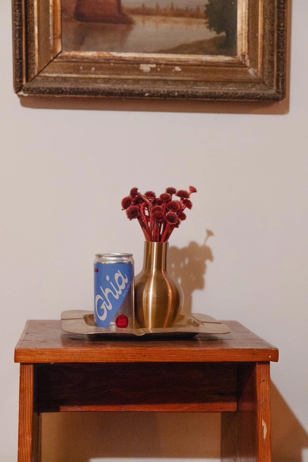 A small wooden table with a brass vase holding red flowers and a flipped blue soda can on a tray, with a framed painting hanging on the wall above.