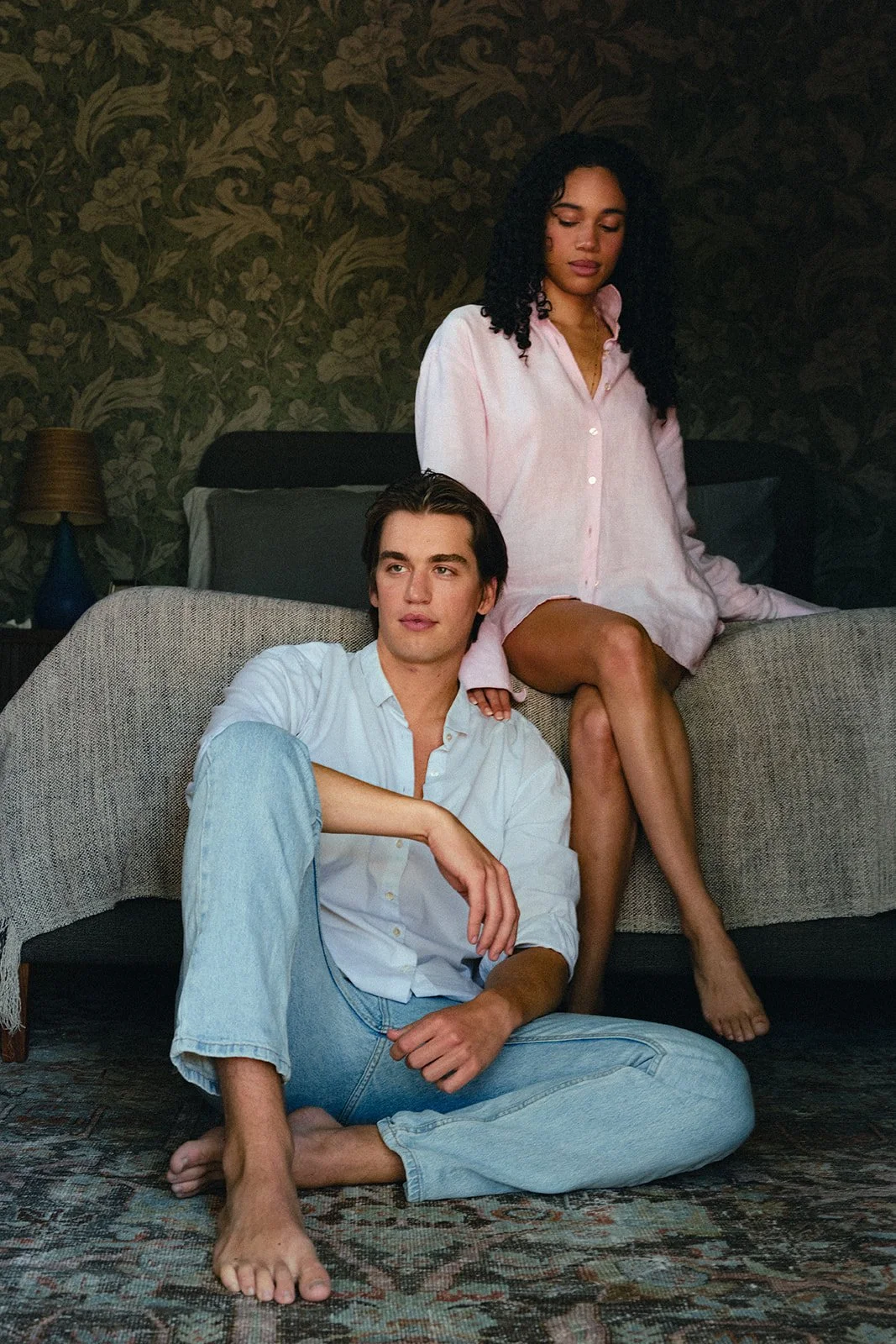 A young man with brown hair, wearing a white shirt and light blue jeans, sitting on the floor with his legs crossed, and a young woman with curly dark hair, wearing a pink shirt dress, sitting on a sofa behind him with one leg crossed over the other, both in a room with dark floral wallpaper and a patterned rug.