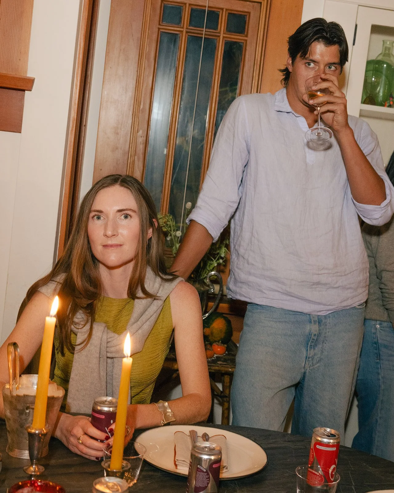 A woman sitting at a table with lit candles, holding a soda can, with candles and dishes in front of her, and a man standing behind her drinking from a glass at a gathering or party.