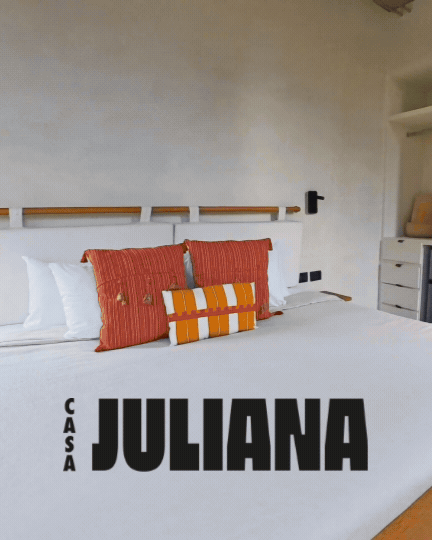A neatly made bed with white linens, two orange and red striped pillows, and a smaller cylindrical pillow with orange, white, and yellow stripes. There are white shelves on the wall, a black wall-mounted lamp, and a dresser with a yellow object in the corner. The text overlay reads "Casa Juliana" in bold black letters.