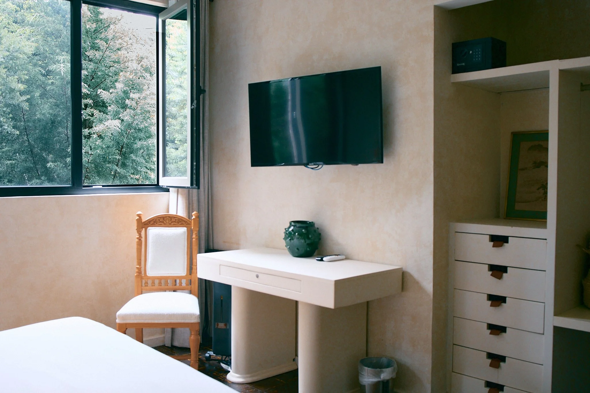 A room with an open window showing green trees outside. Inside, there's a wall-mounted flat-screen TV, a small white desk with a green vase and a remote control, a white chair with a wooden frame, a tall white dresser with drawers, and a small safe o