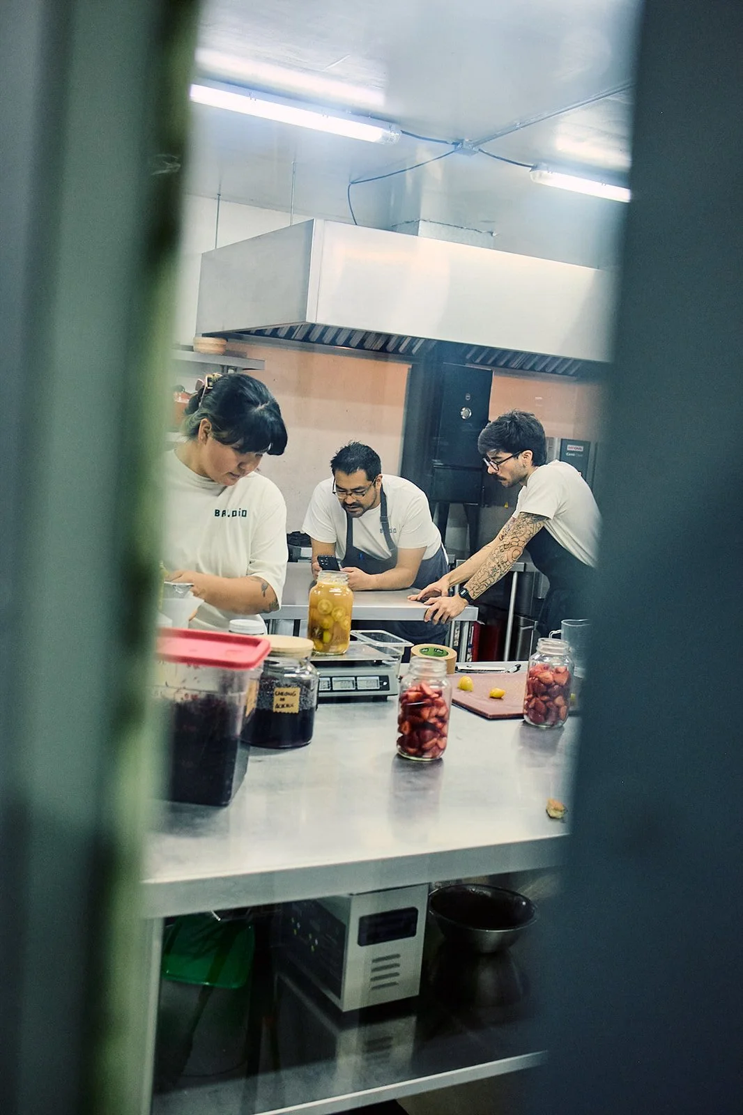 Three people in a commercial kitchen preparing food, viewed through a small opening or gap in a door or partition