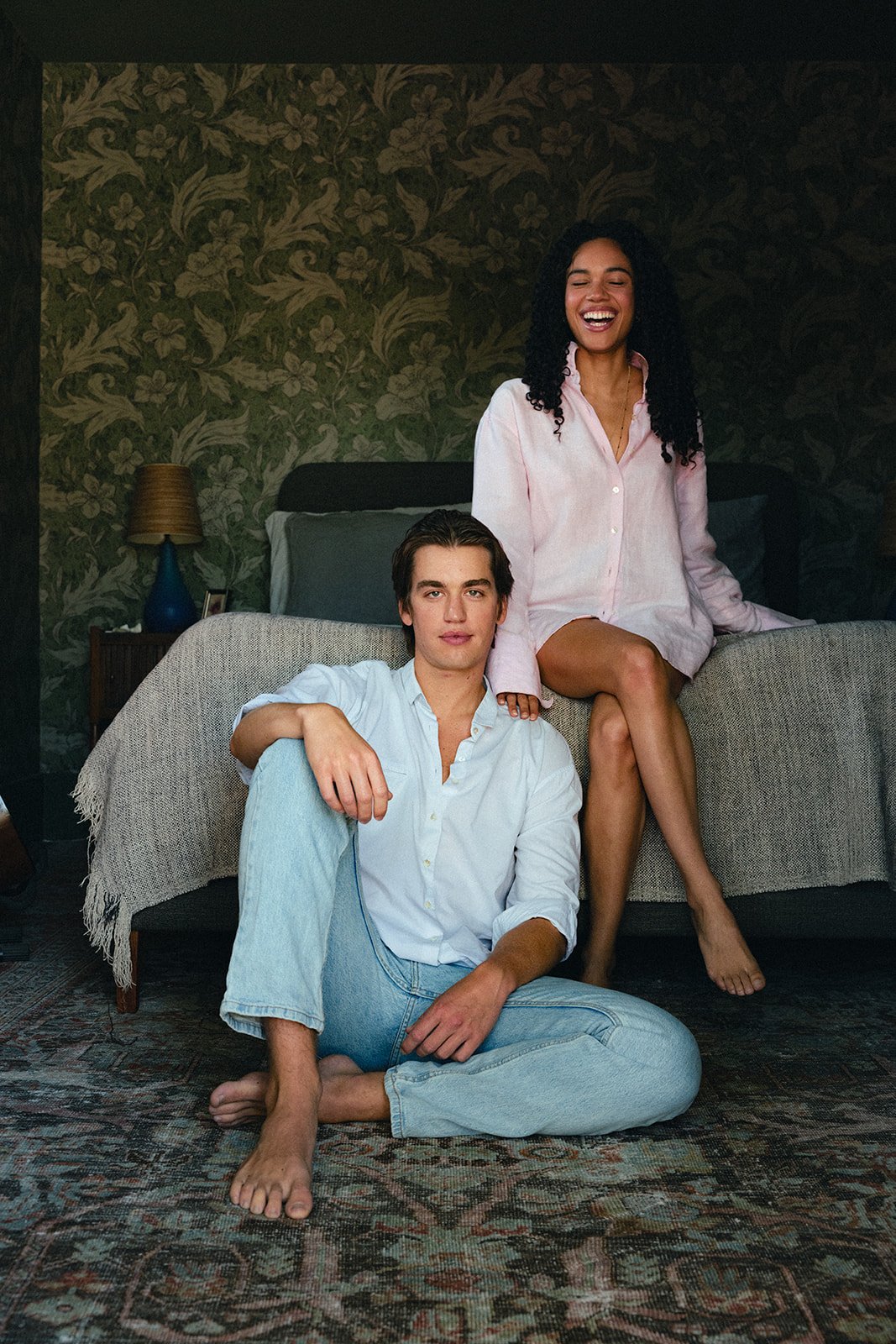 A young man and woman in a bedroom, with the woman sitting on a bed and the man sitting on the floor in front of her. Both are smiling and relaxed. The bedroom has floral patterned wallpaper, a bedside table with a lamp, and a blanket on the bed.