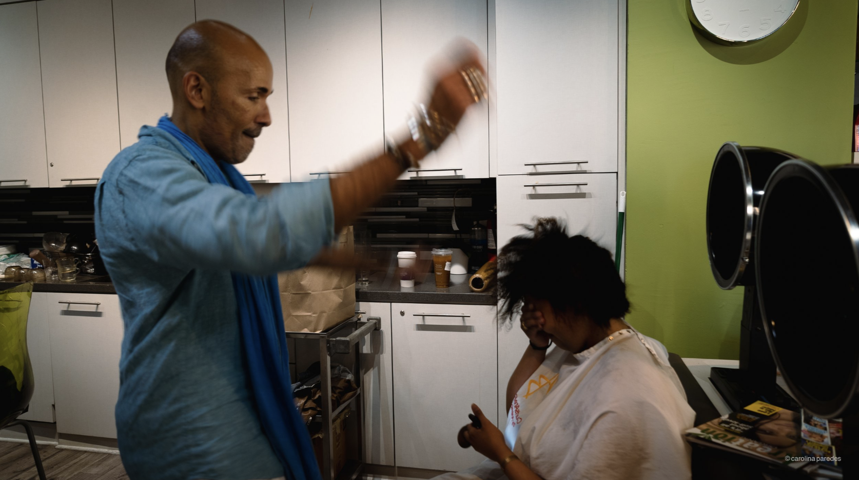 A man with a shaved head and wearing a blue scarf raises his hand as a woman with messy hair covering her face rests her head on her hand in a hospital or clinic room.