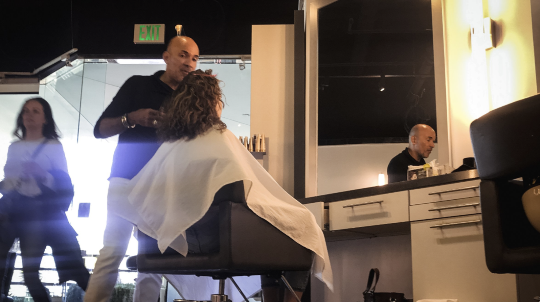 A woman with curly hair sits in a salon chair with a white cape. A hairstylist stands nearby. In the background, a man sits at the salon counter and a woman walks past. The salon has a black and white color scheme with modern decor.