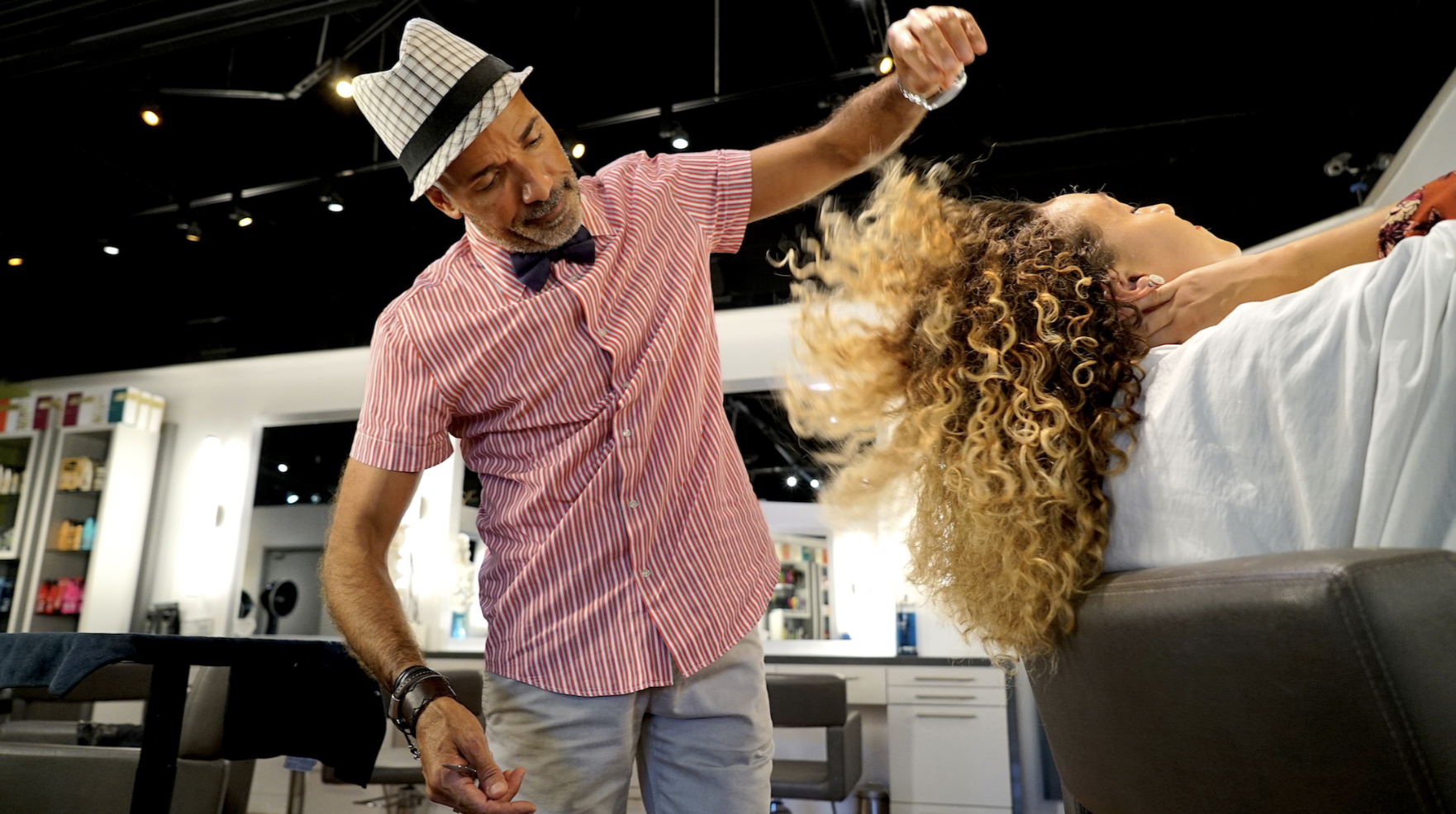 A man with a checked fedora hat, striped red shirt, and bow tie is haircutting a woman's curly hair in a salon.