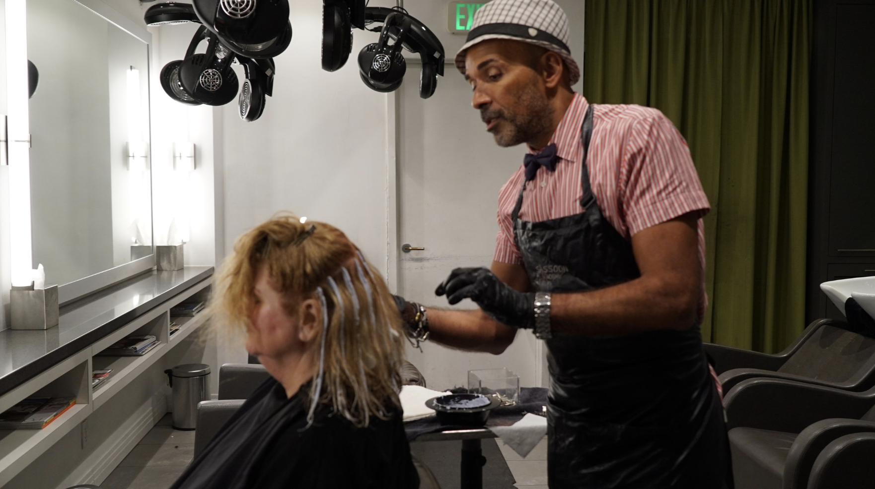 A man with a checkered hat and striped shirt is dyeing a woman's hair in a salon. The woman has curly, blonde hair and is seated in front of a mirror. The salon has hairdryers hanging from the ceiling, a large mirror, and green curtains in the backgr