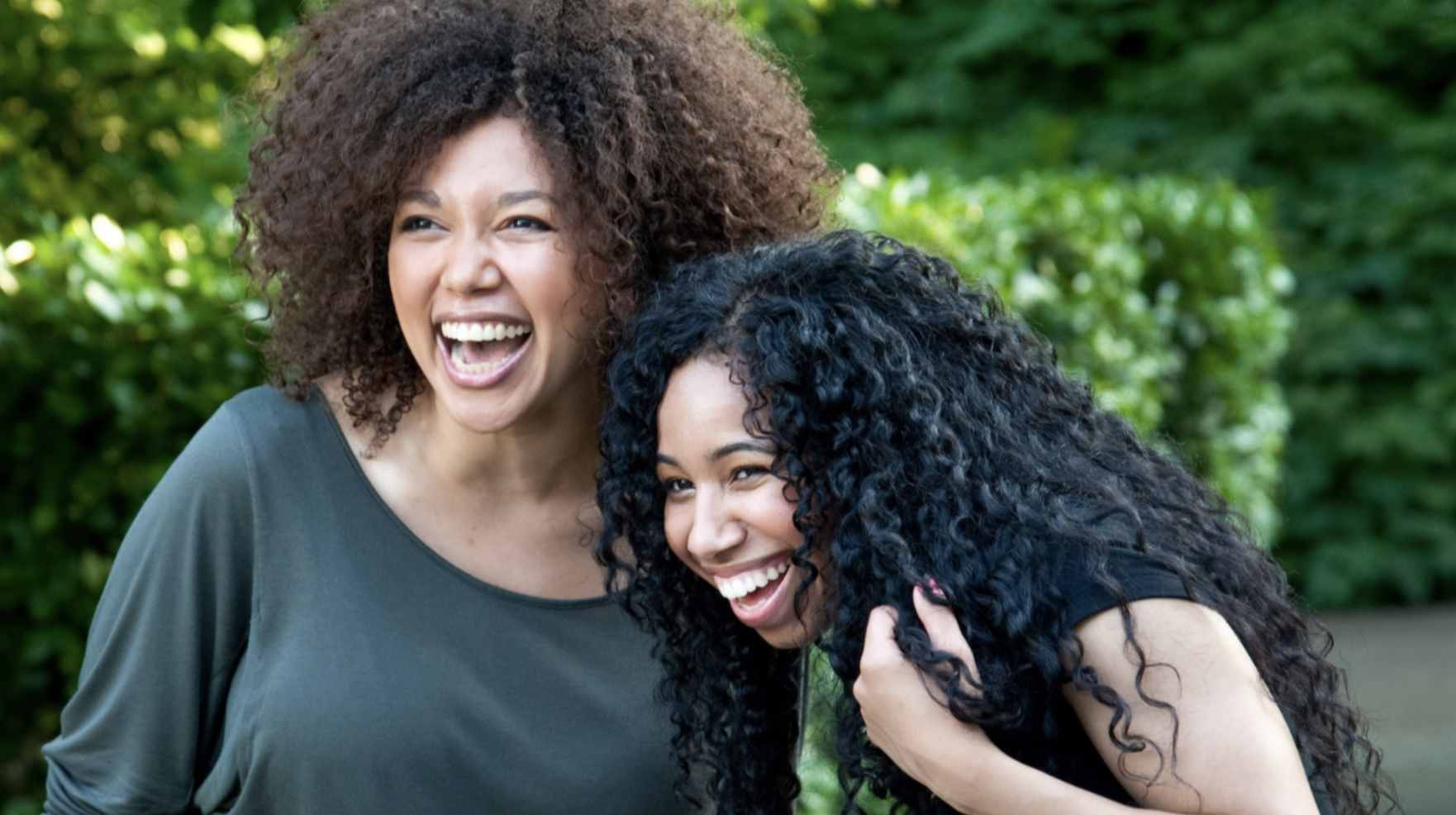 Two women with curly hair laughing together outdoors in a green park.