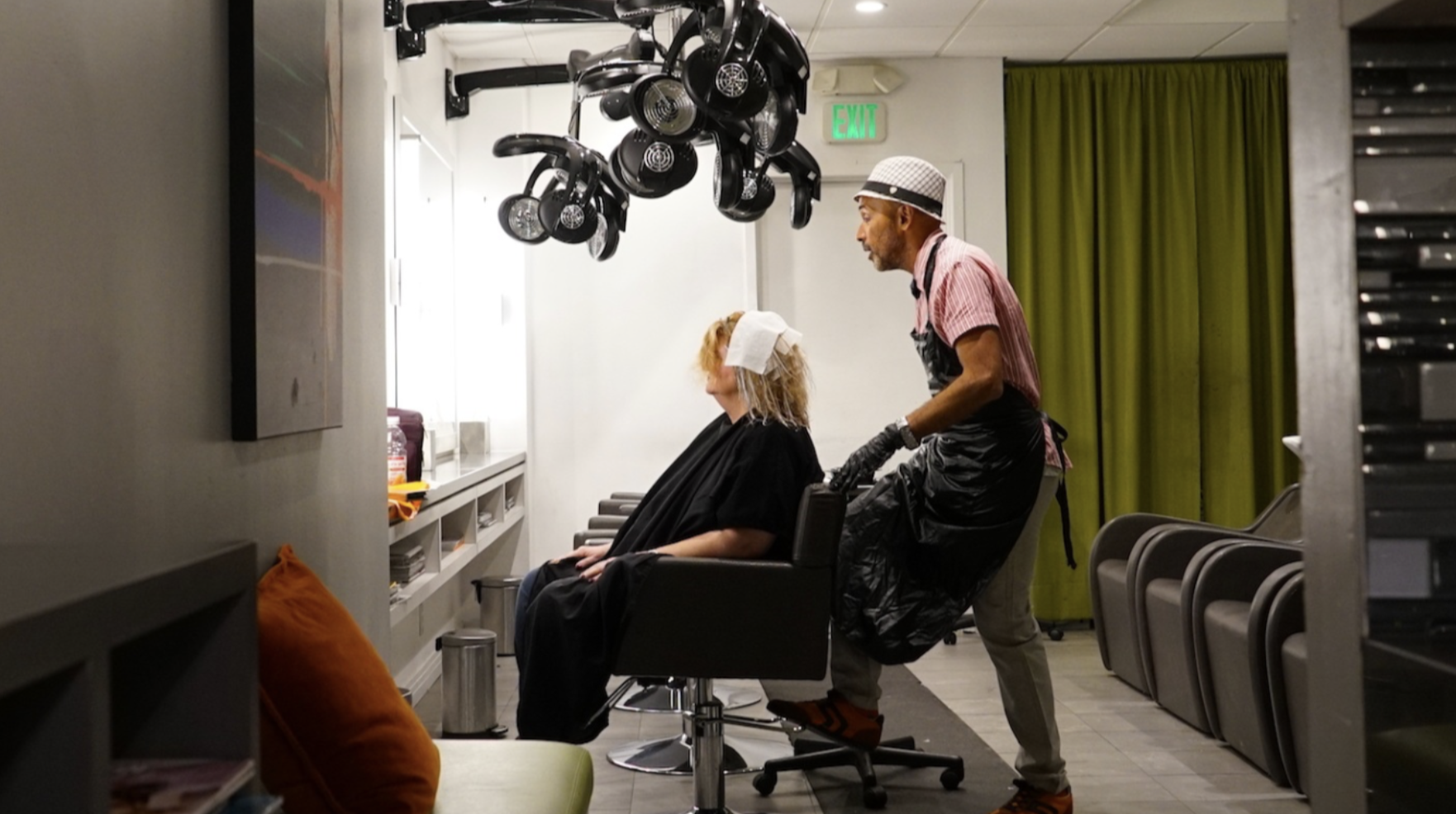 A hairstylist in a pink striped shirt, black apron, and white hat is working on a woman's hair at a salon. The woman, with curly blonde hair and a towel on her head, is seated in a black salon chair. Hairdryers hang from the ceiling behind them, and