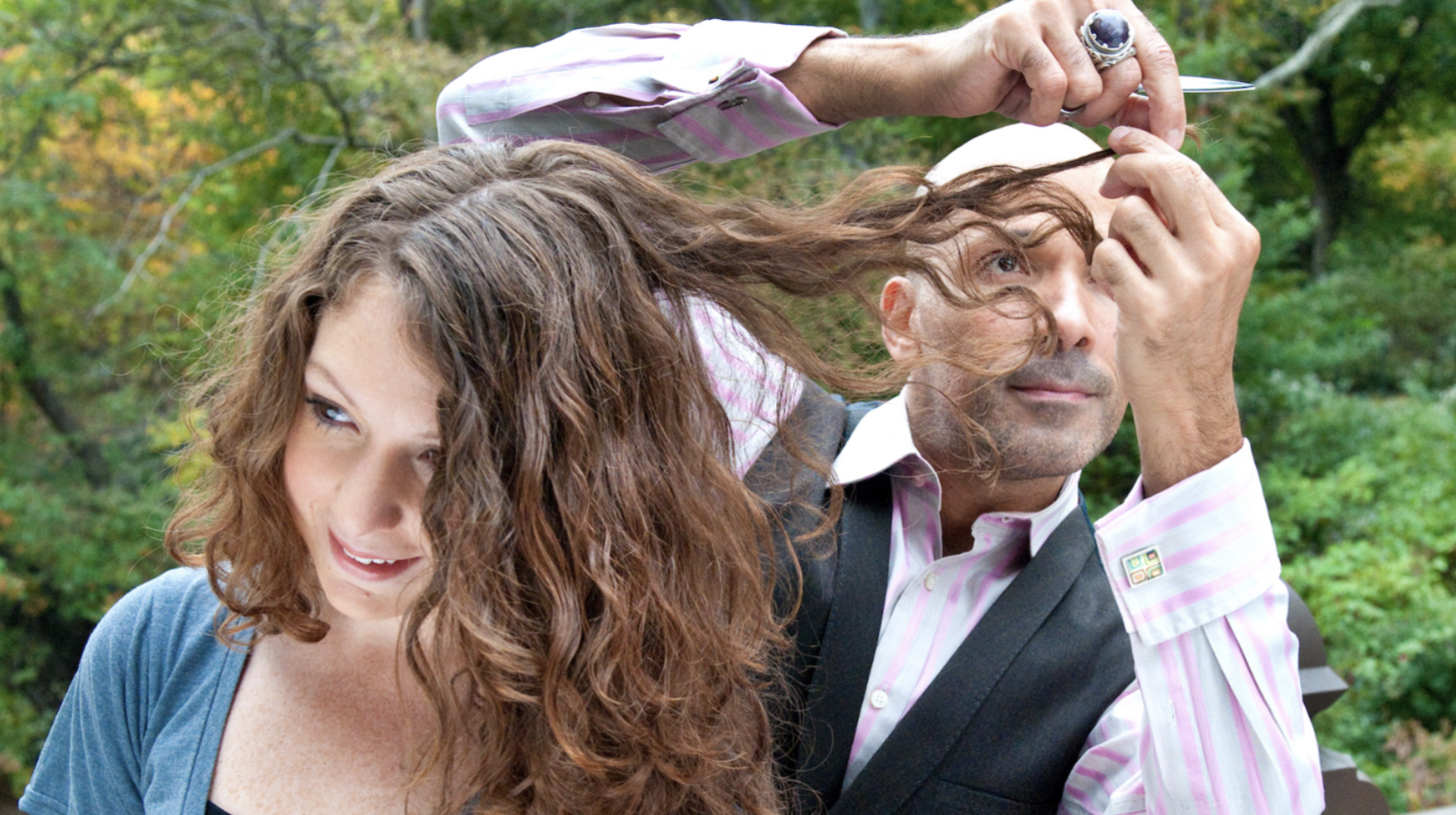 A man and a woman outdoors, the man is holding a small knife and a lock of the woman's hair, the woman is smiling, and green trees are in the background.