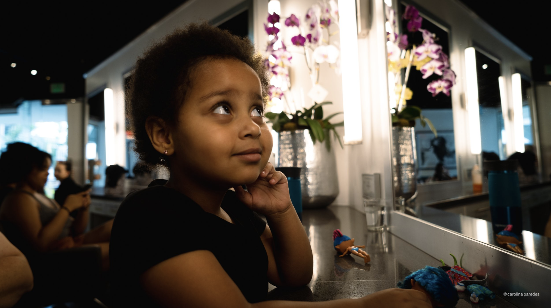A young girl with curly hair and earrings sits at a counter in a salon, looking upwards with a thoughtful expression. There are orchids in vases, mirrors, and other people in the background, some using their phones.