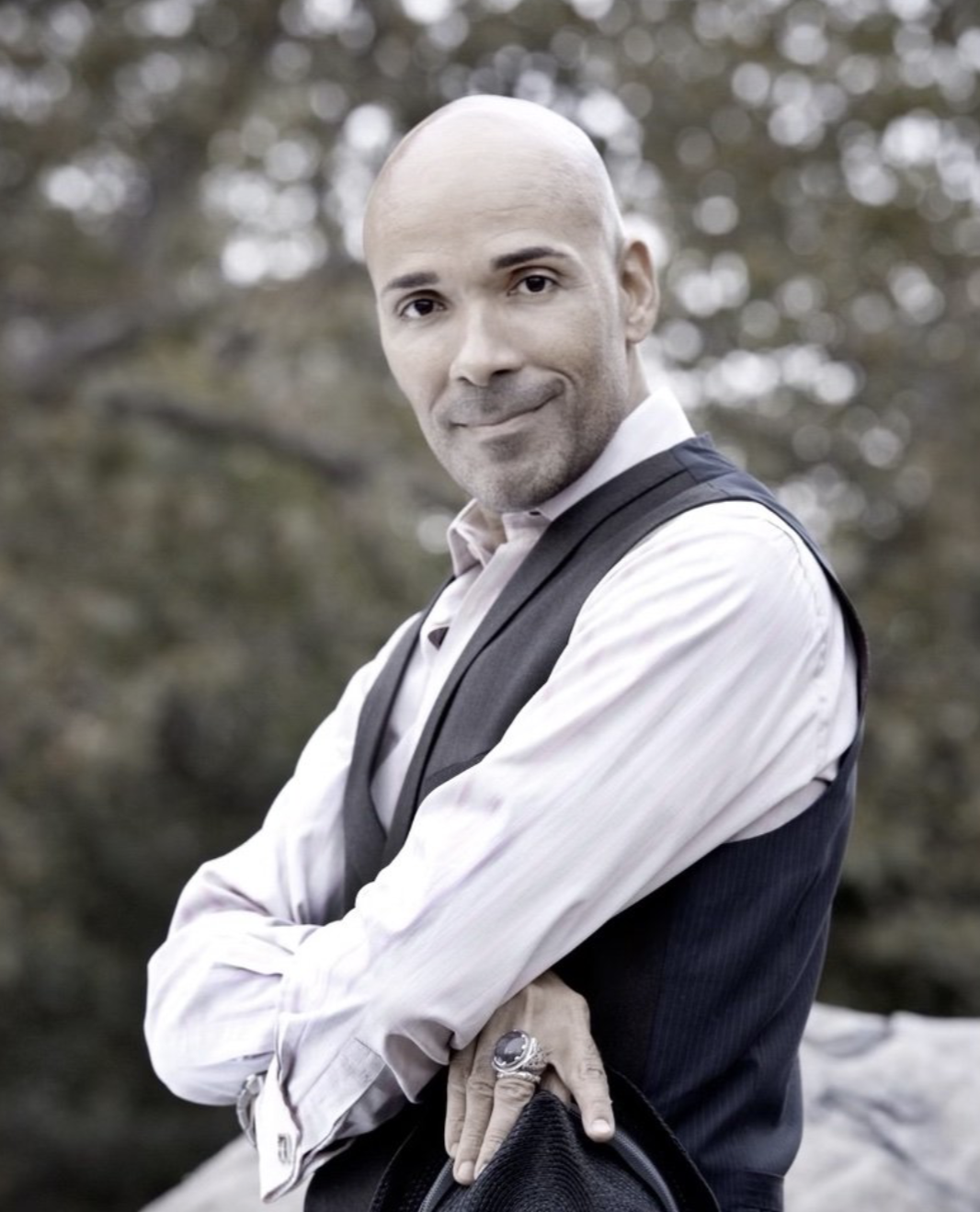 A bald man wearing a light-colored shirt and a dark vest, standing outdoors with trees in the background, smiling slightly and crossing his arms.