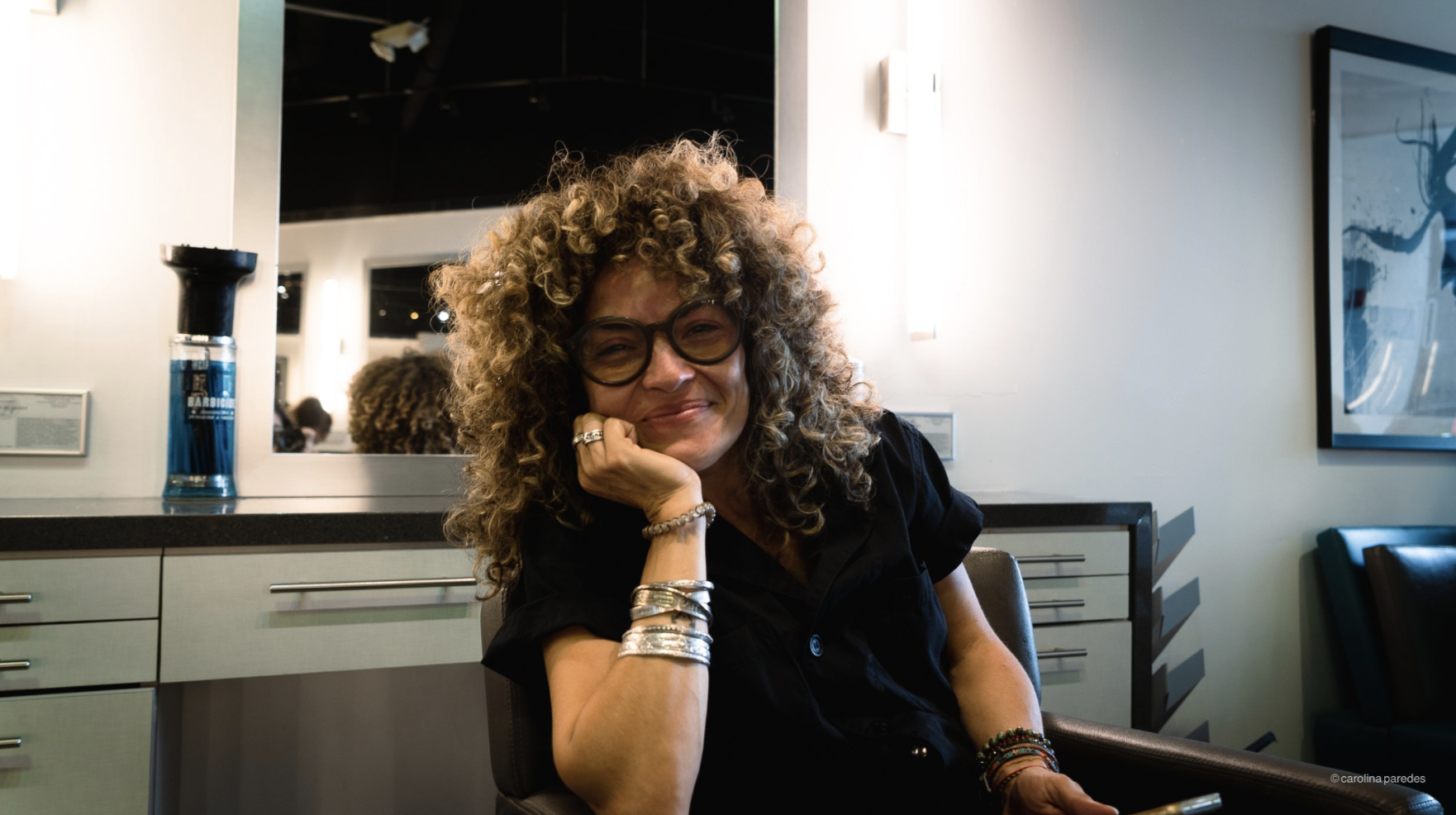 A woman with curly blonde hair and glasses smiling, sitting indoors with her chin resting on her hand. She is wearing black clothing and multiple bracelets.