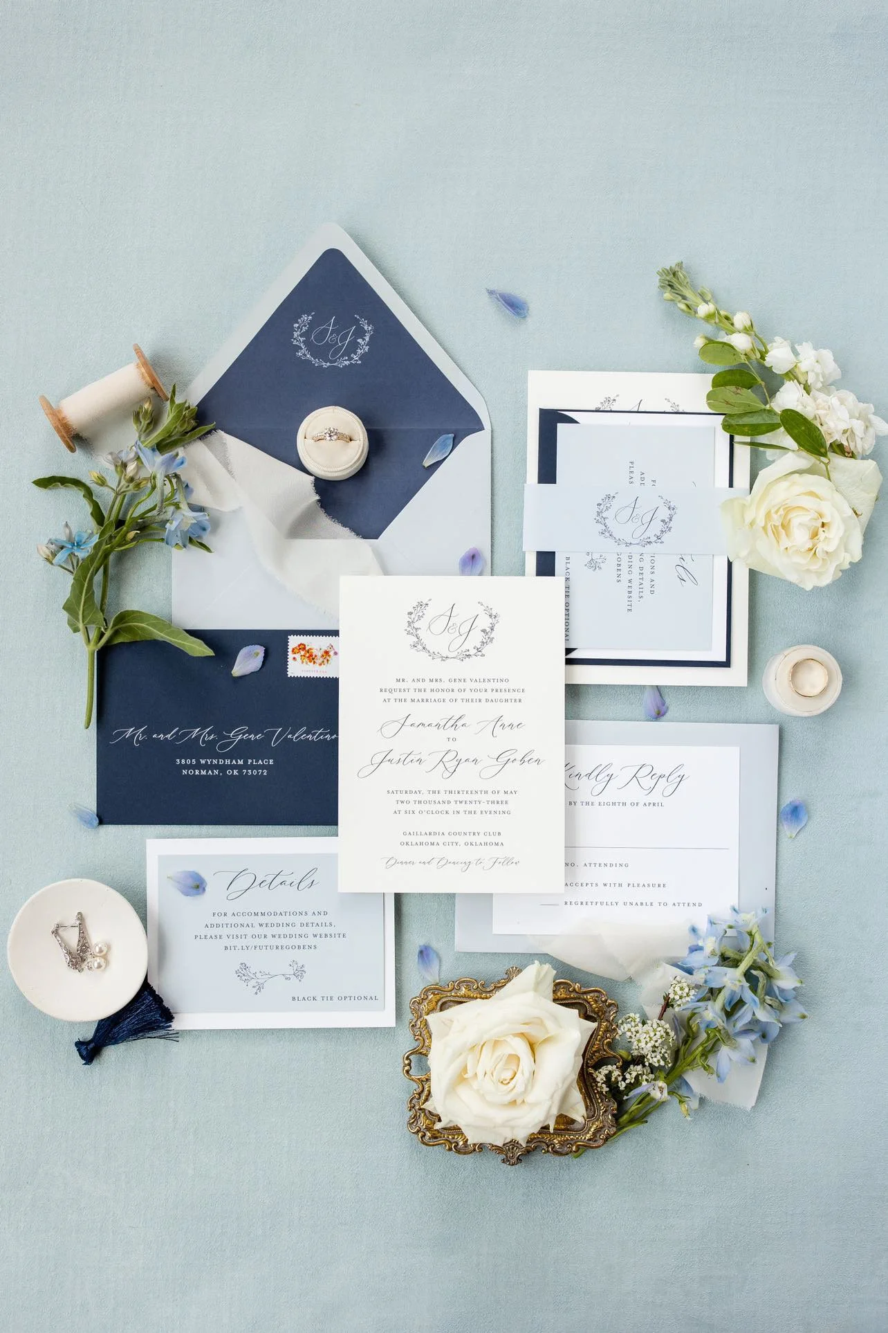 Elegant wedding invitation suite with navy and white color scheme, floral accents, wedding rings, and small decorative items on a light blue background.