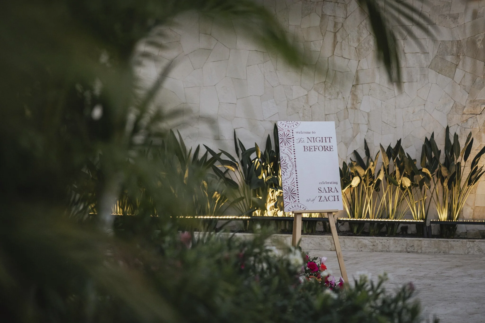 Wedding reception sign reading 'welcome to the night before celebrating Sara and Zach' amidst decorative plants and flowers with a stone wall background.