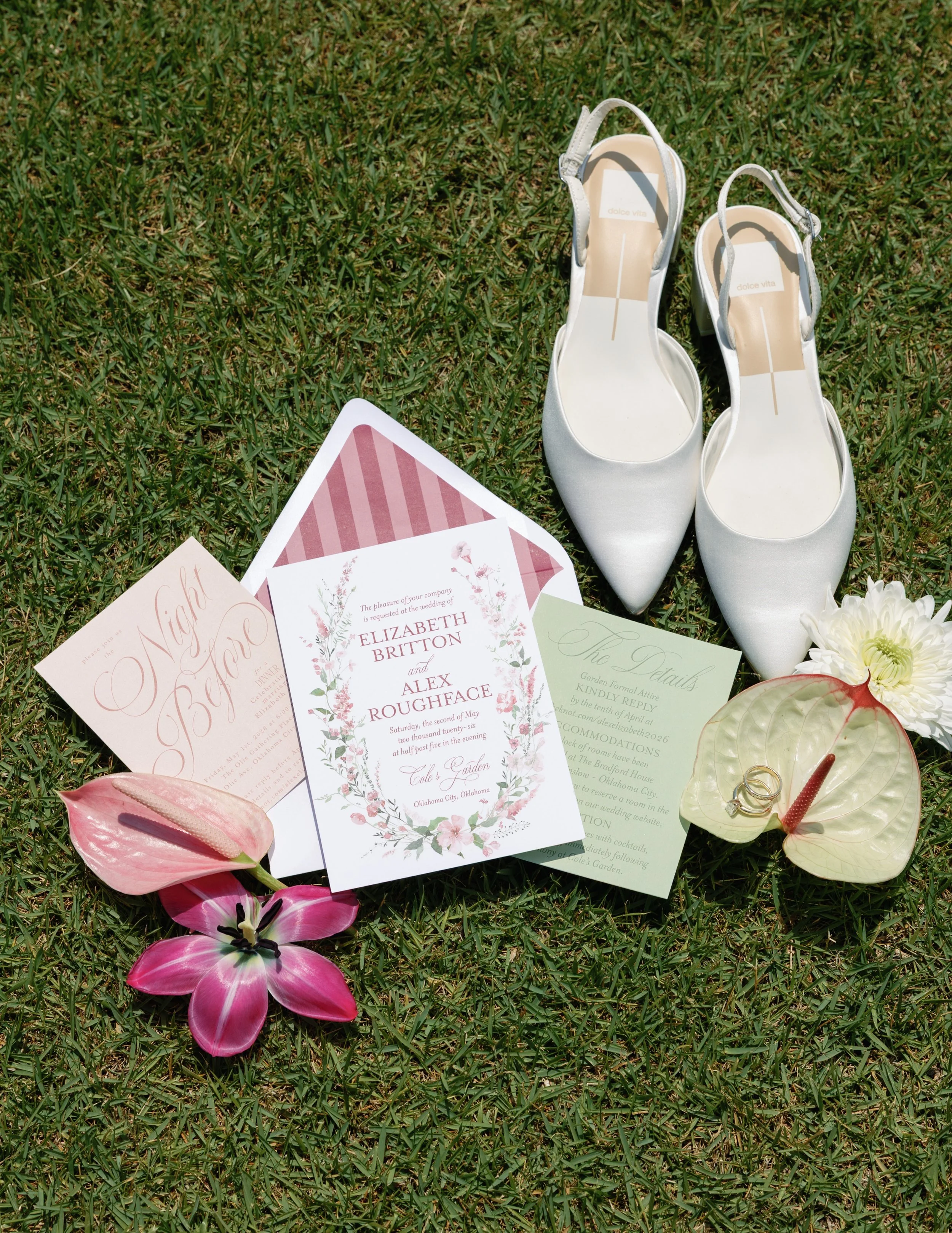 A pair of white high-heeled shoes, wedding invitations, pink and white flowers, and a glass dish with two rings on grass.