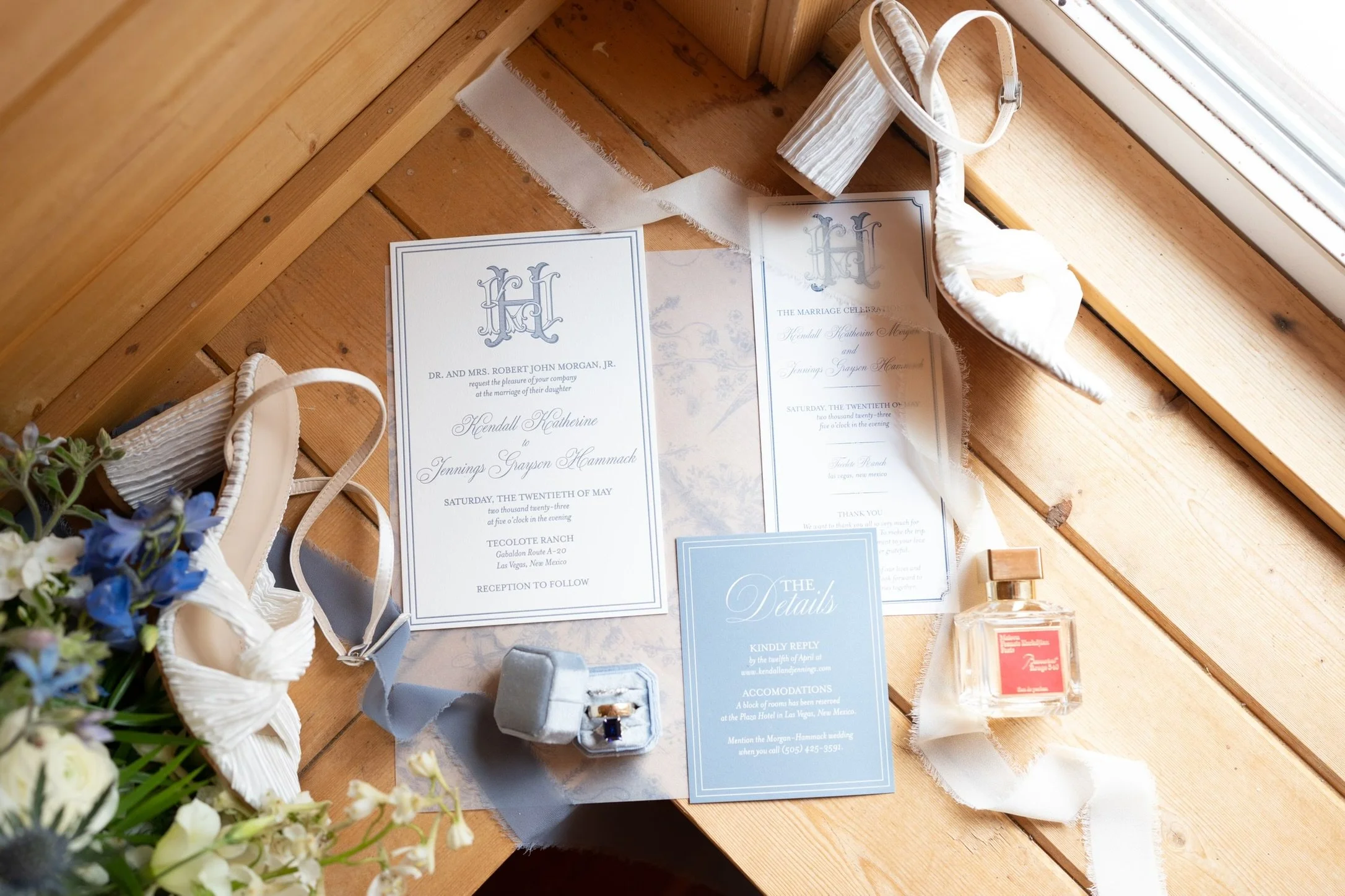 Wedding invitation cards, a small jewelry box with a ring, and floral arrangements on a wooden table.