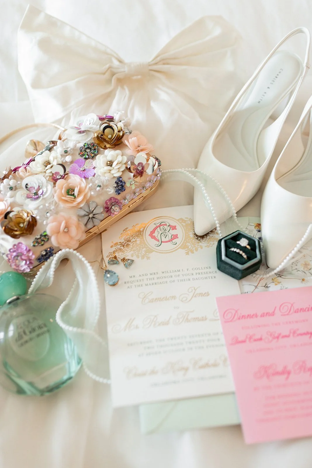 Collection of elegant wedding accessories including a pair of white high-heeled shoes, a floral decorated clutch, pearl jewelry, a wedding invitation, perfume bottle, earrings, and a small ring box.