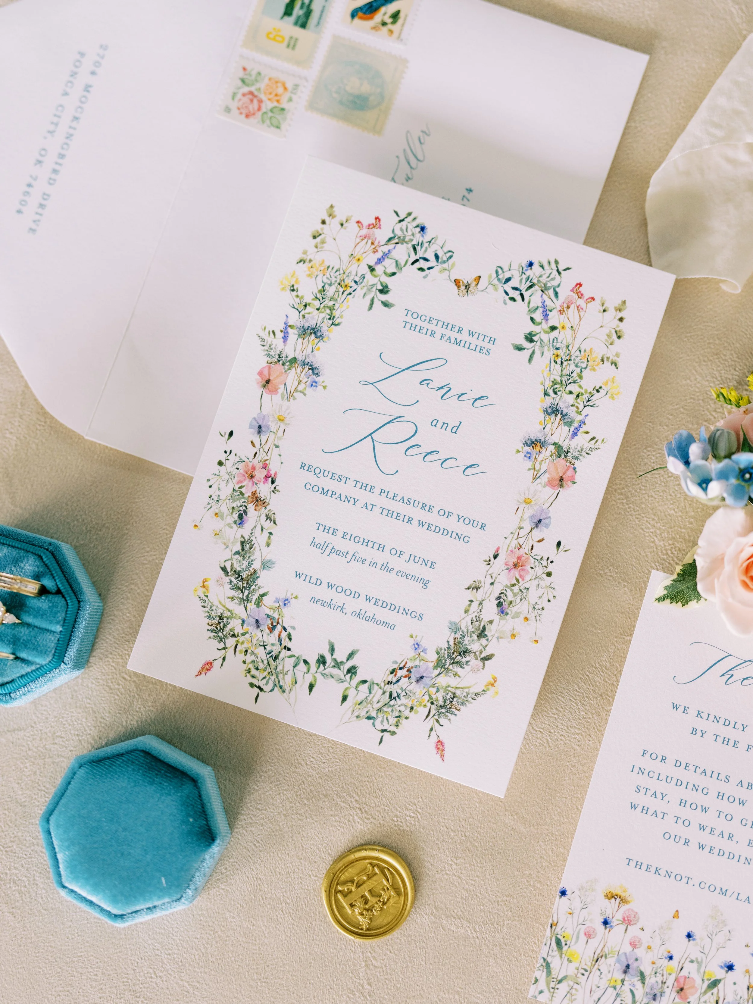 A wedding invitation with floral border design, including a butterfly, surrounded by wedding-related items such as a gold wax seal, turquoise ring box, and postal stamps.