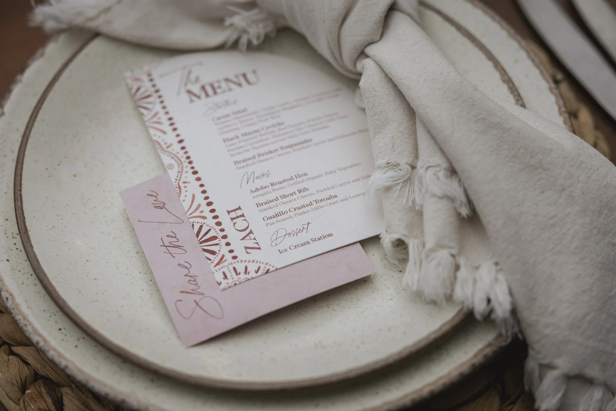 A menu card placed inside a beige fabric cover, which is partially laid on a speckled ceramic plate. The menu is decorated with red accents and includes listed food items. The plate is on a woven basket surface.