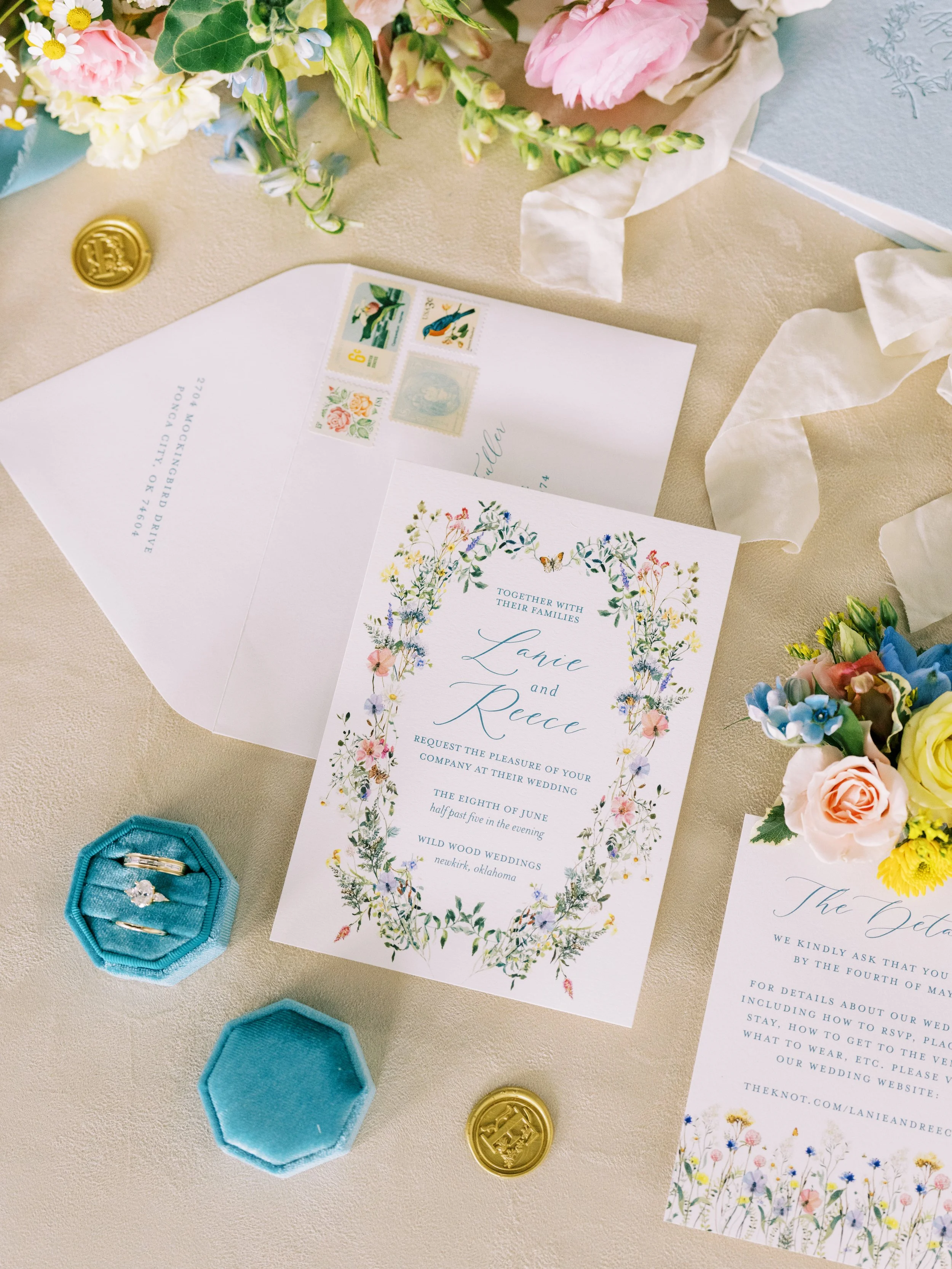 Wedding invitation with floral border and names Lanie and Reece, surrounded by pink and yellow flowers, gold wedding seals, and blue ring boxes containing rings.