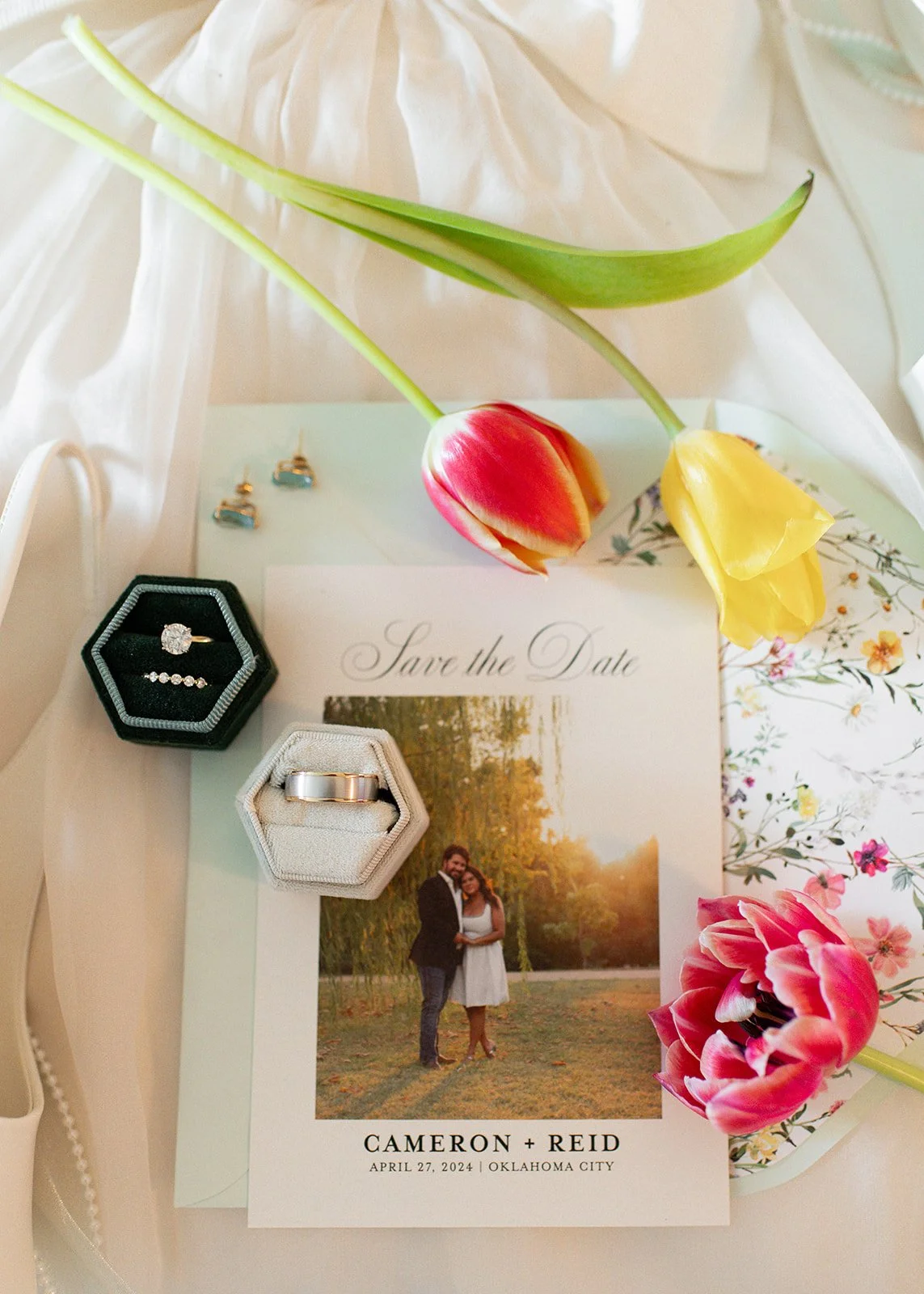 Wedding save the date card with a photo of a couple, pink and yellow tulips, a pair of earrings, a wedding ring in a black box, and a wedding band in a white box, all on a white surface with floral and floral patterned elements.