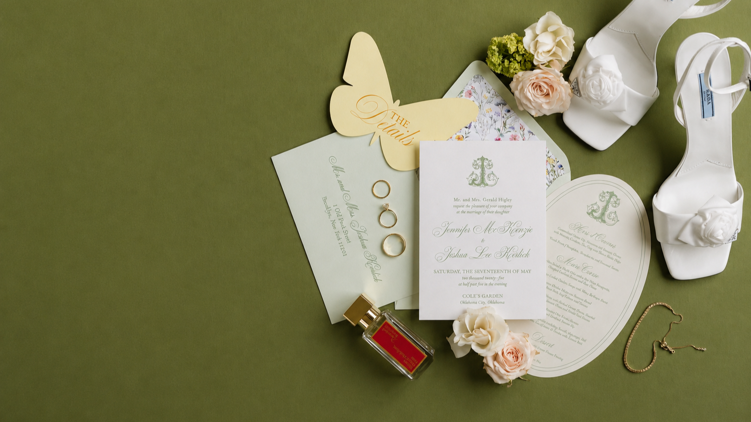 Wedding invitation with flowers, shoes, jewelry, and perfume on a green surface.