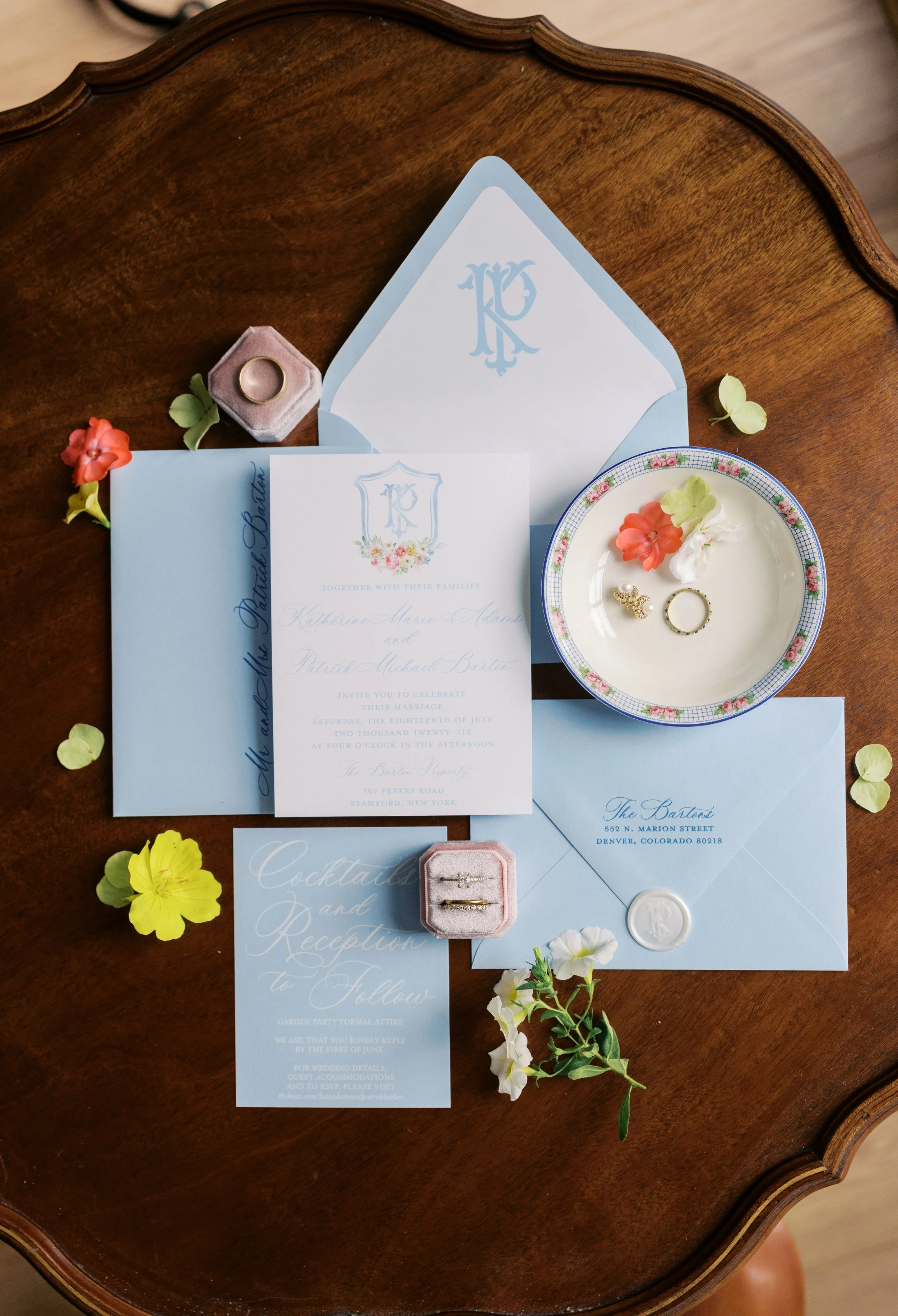 Wedding invitation suite with light blue envelopes, floral graphics, and gold rings in a pink ring box on a wooden table, accompanied by flowers.