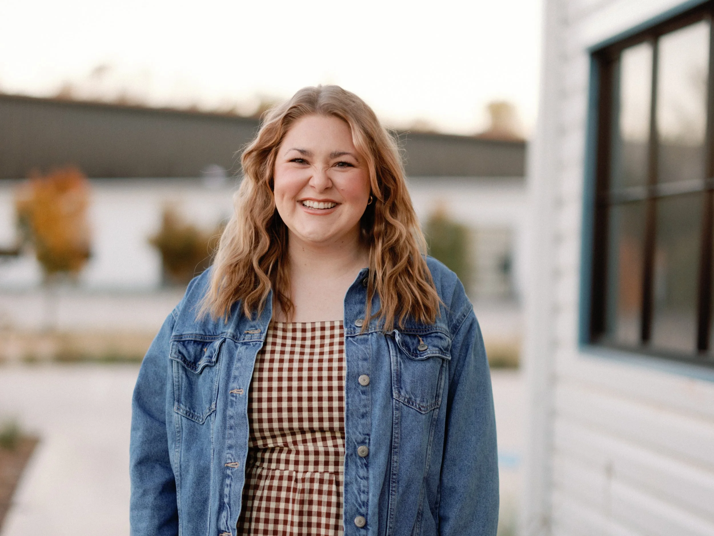 Young woman with long curly hair smiling outdoors, wearing a denim jacket over a checkered shirt.