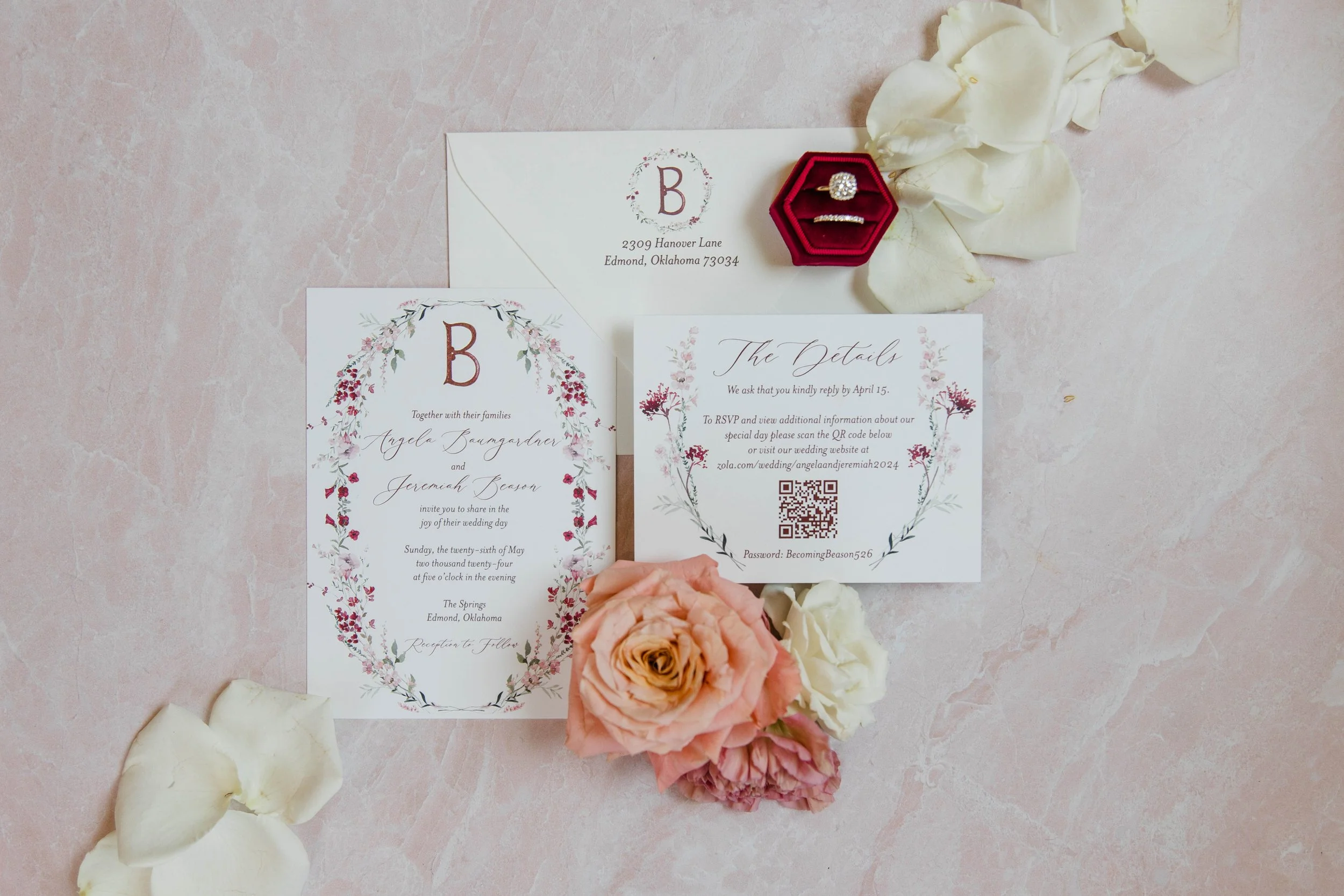 An invitation and RSVP card for a wedding, with floral designs and a QR code, surrounded by cream and peach roses and white petals on a pink marble surface.