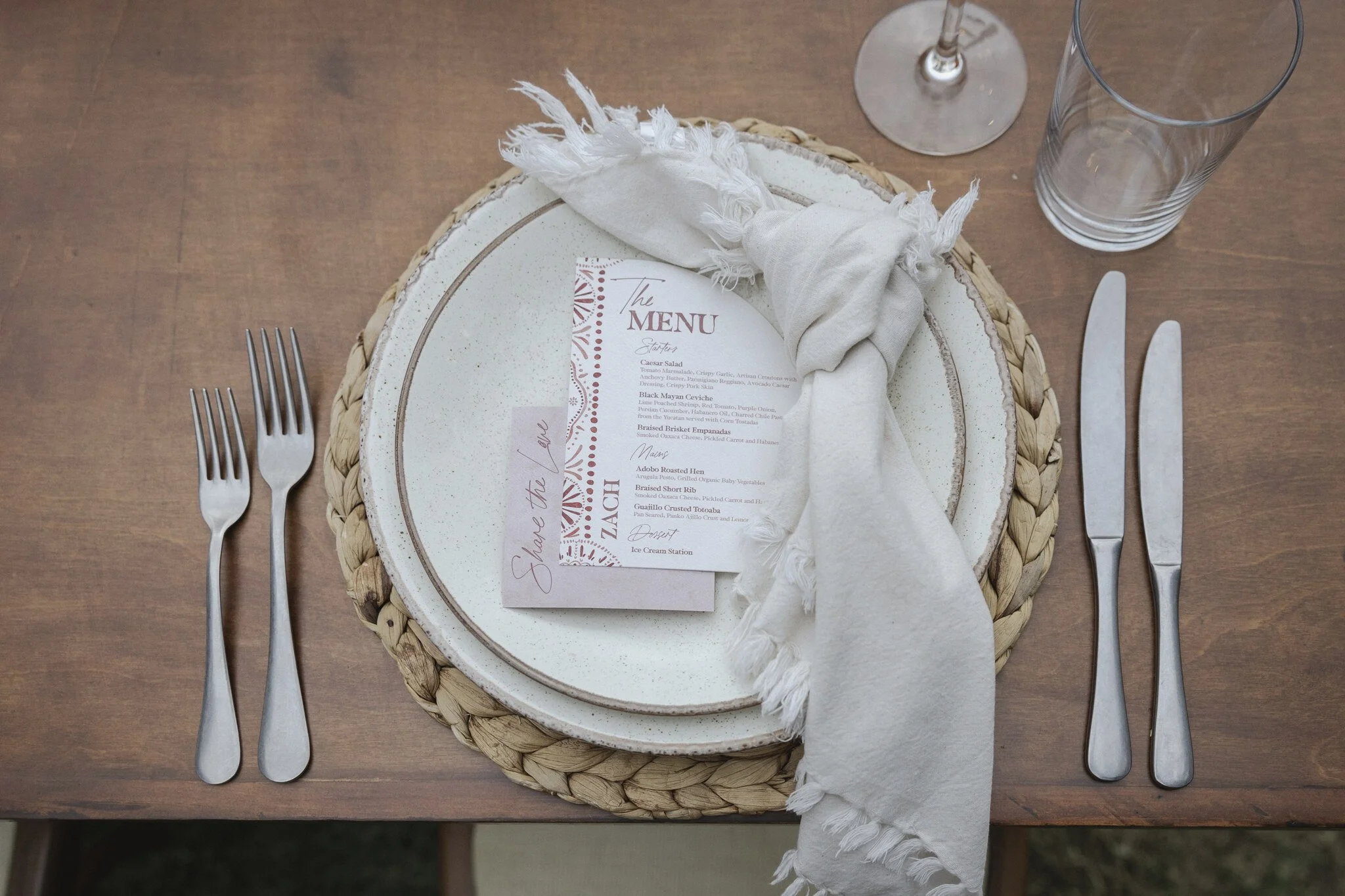 Table setting with placemat, white cloth napkin, menu card, forks, knife, empty glass, and wine glass on a wooden table.