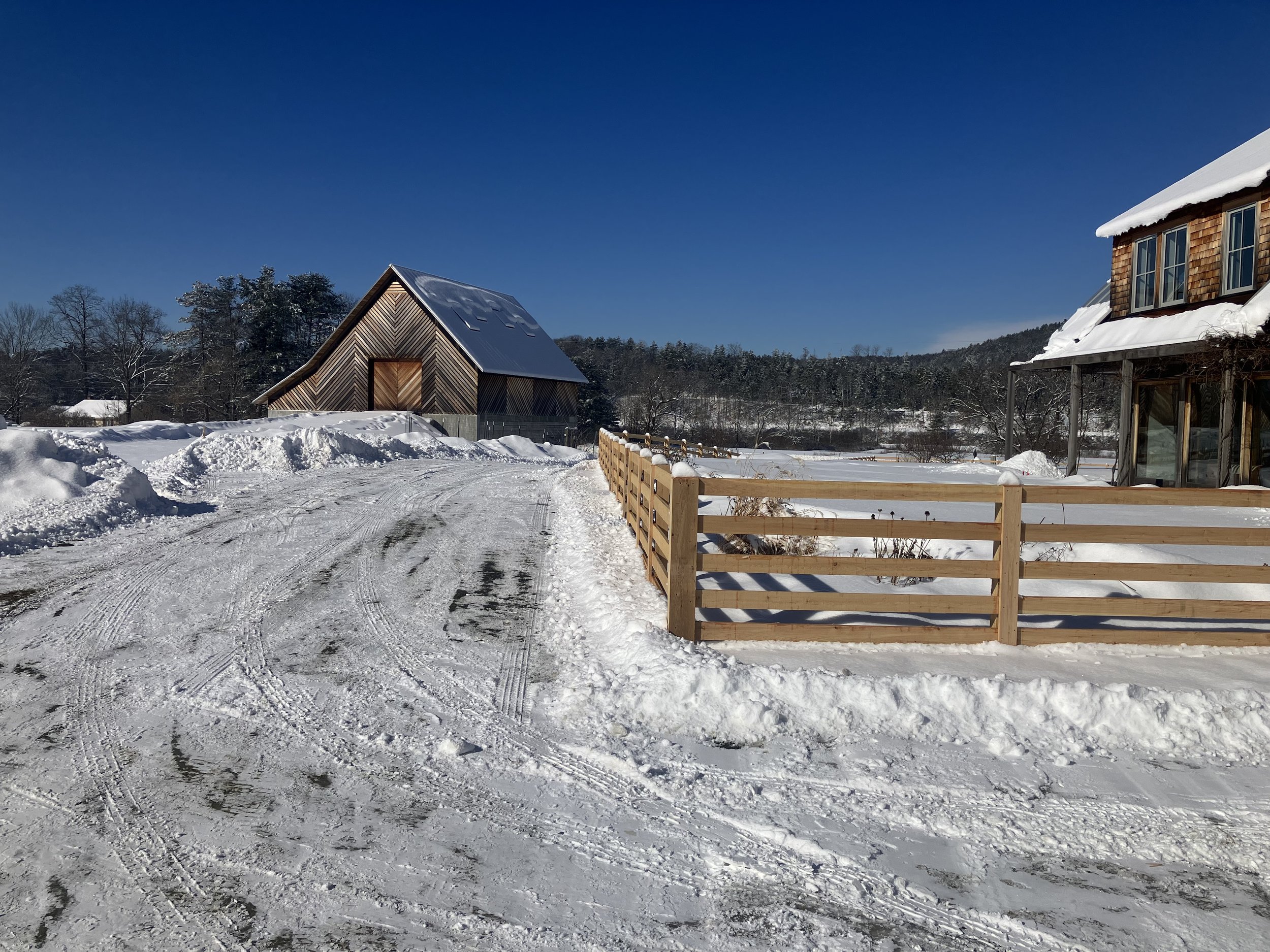 Snowy rural landscape with a fence, a modern wooden barn, and a house under a clear blue sky.