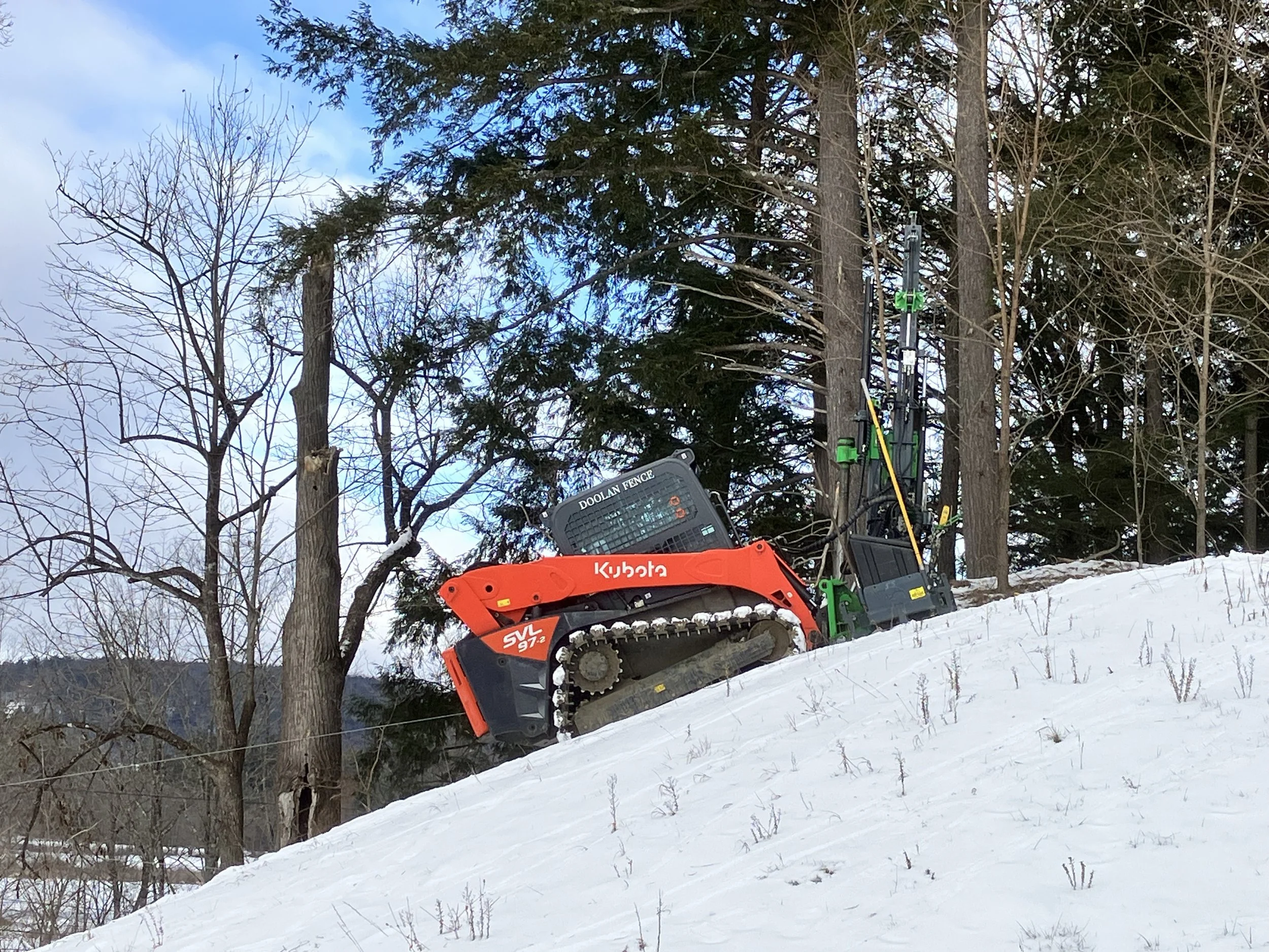 A red Kubota SVL 97-2 compact track loader on a snowy hillside, equipped with forestry attachments, surrounded by trees and a partly cloudy sky.