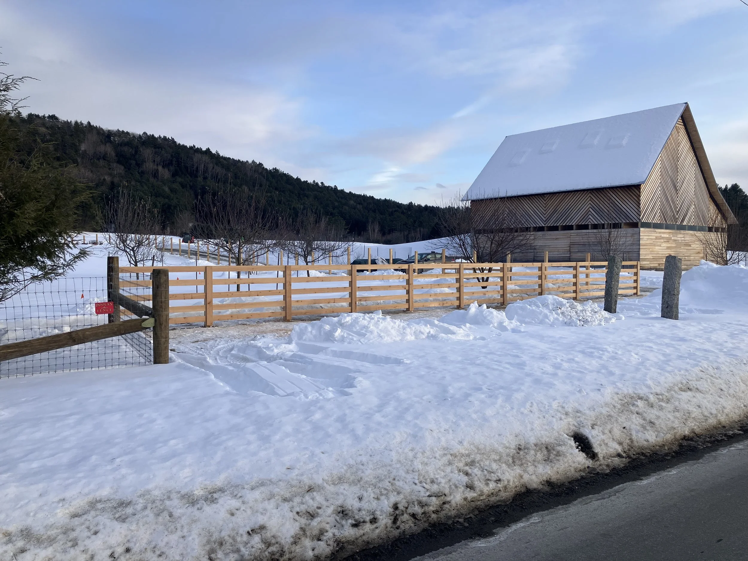 A snow-covered rural scene with a wooden house, a wooden fence, and a forested hillside in the background.