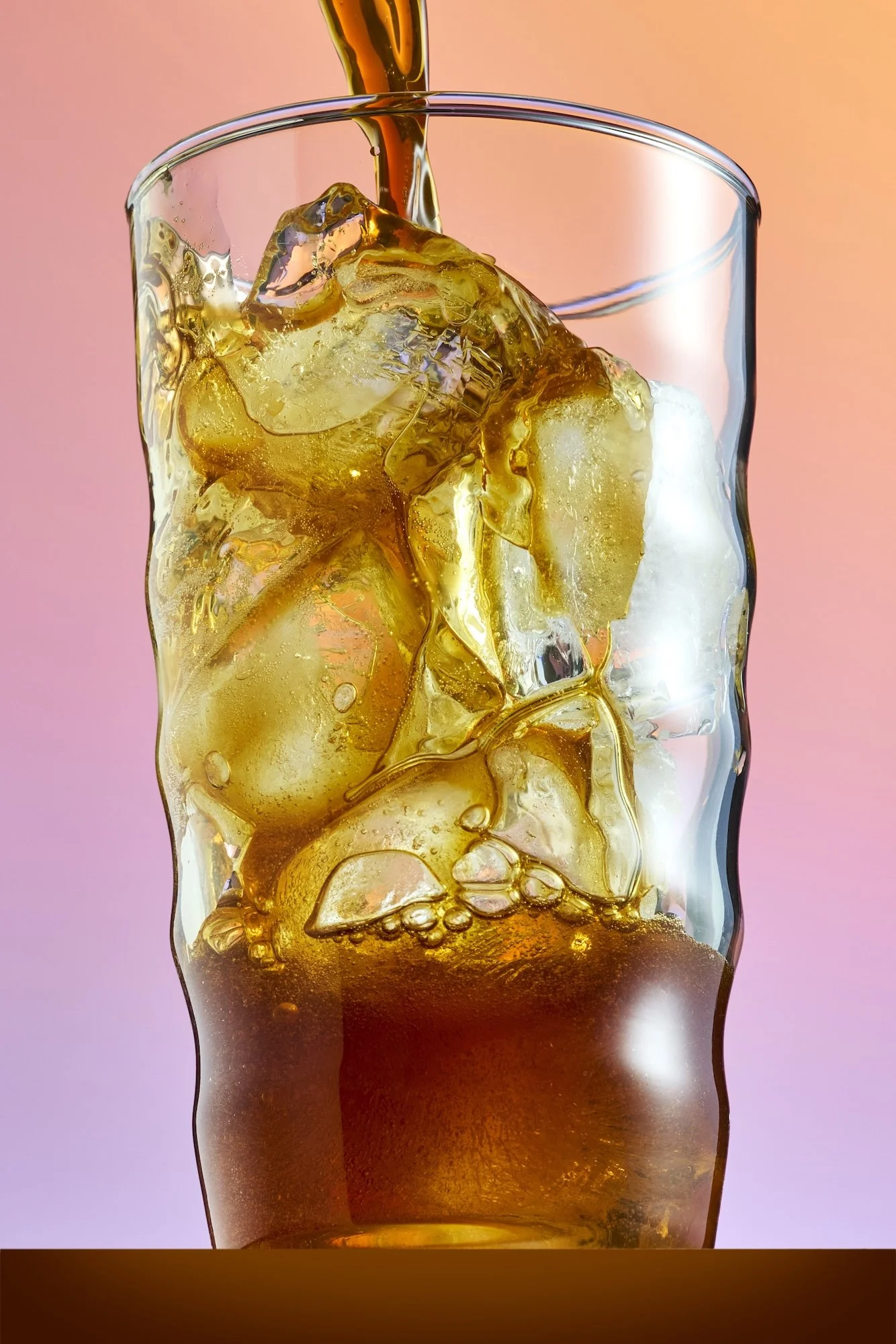 A close-up of a glass filled with ice cubes and cola, with cola being poured into the glass. The background is a gradient of pink and orange hues.