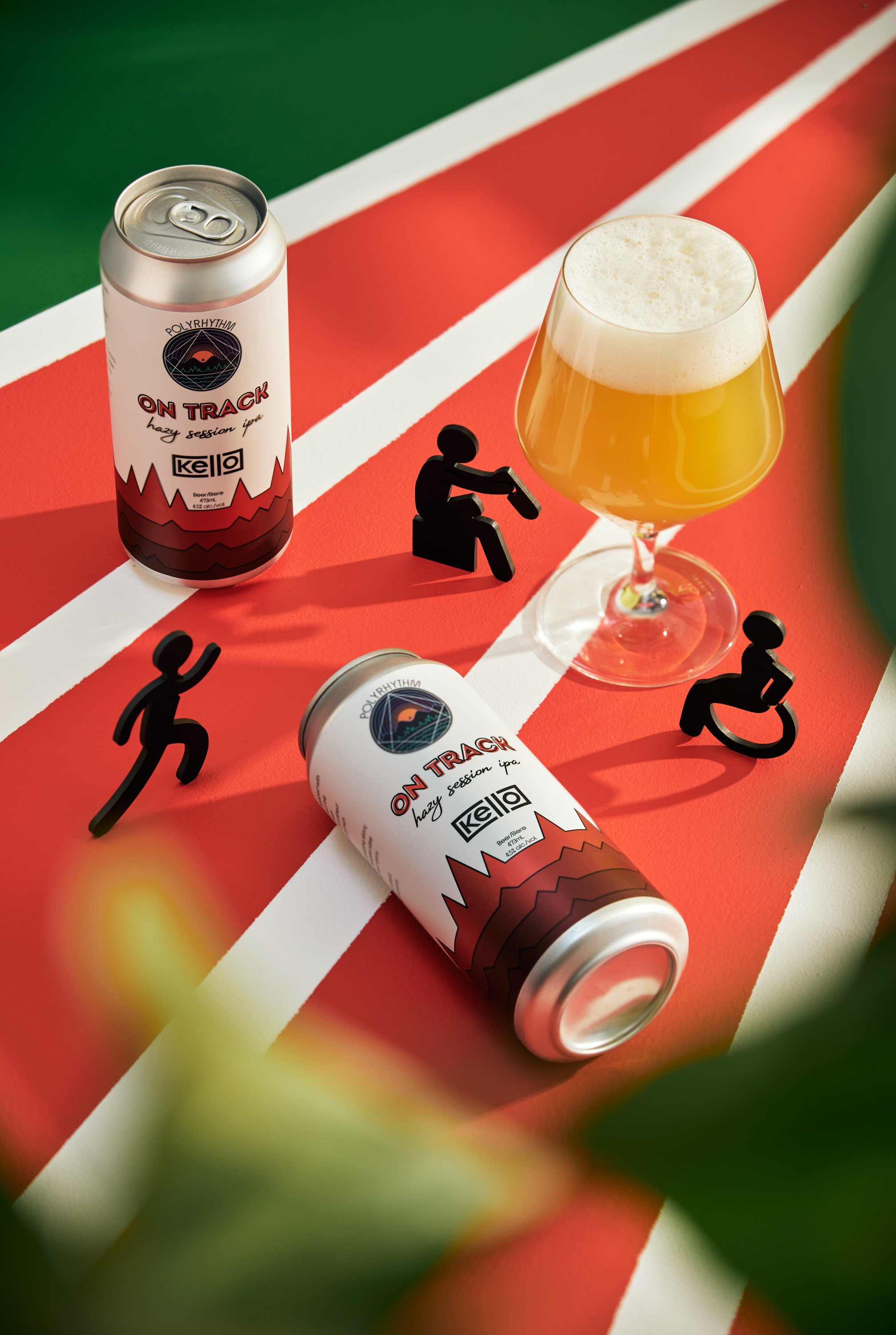 A can of Polly Rhythm On Track hazy session IPA beer, and a glass of beer on a red, white, and green striped surface with small black figurines of people, some in wheelchairs, and a green background.