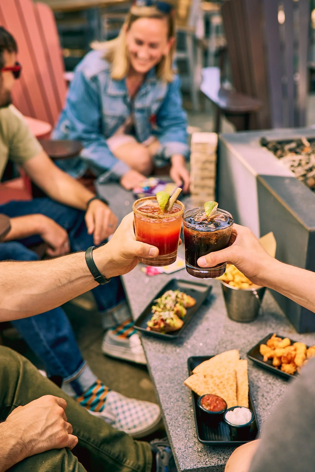 People raising glasses with cocktails at a table filled with snacks and food in a lively social gathering.