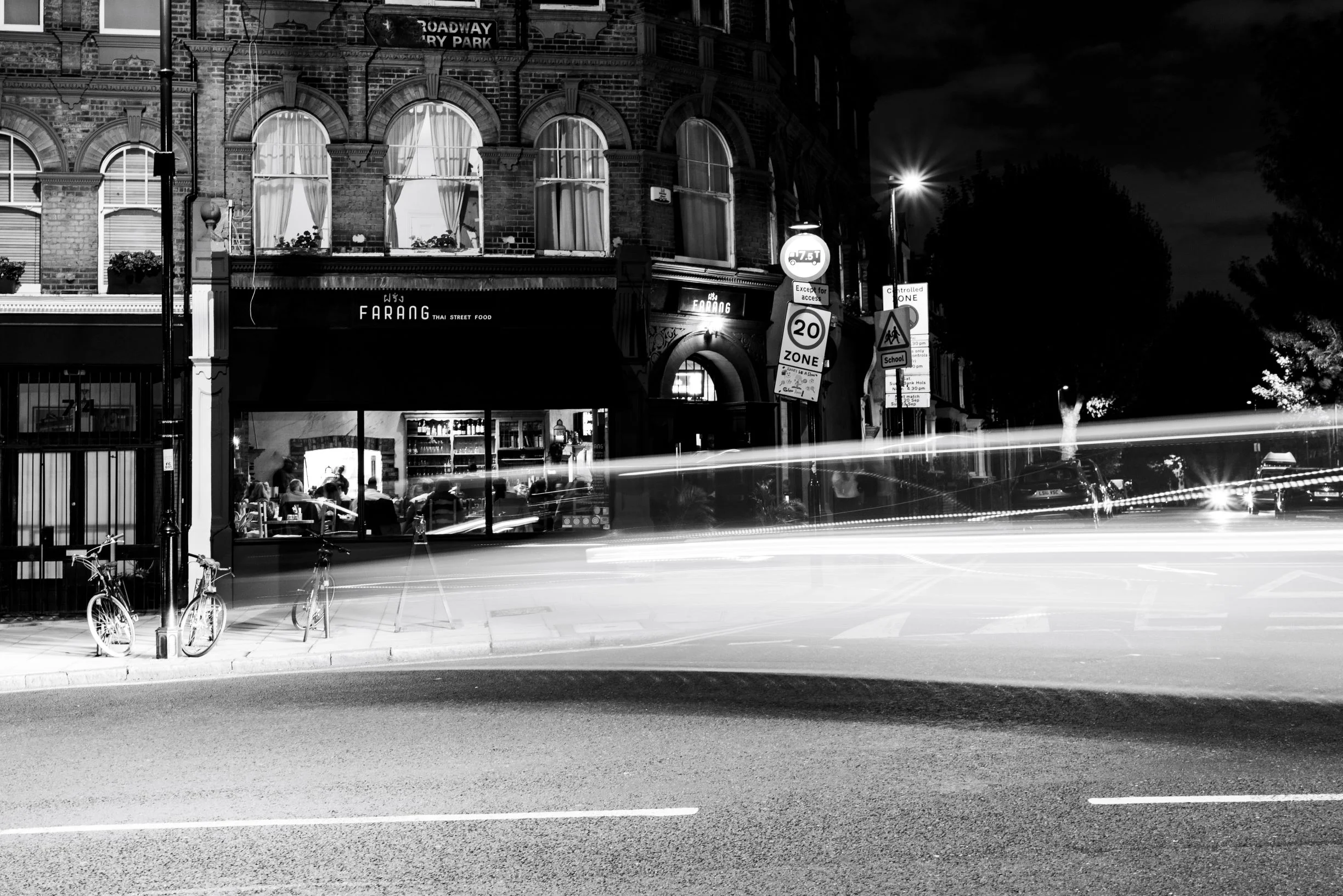 Farang London restaurant exterior at night in black and white, with street lights and traffic light trails on Highbury Park, photographed by Sebby Holmes.