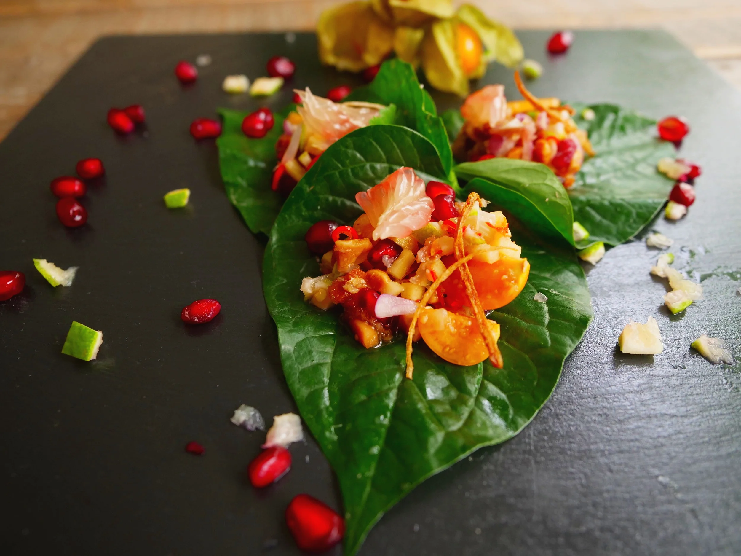 Pomegranate miang bites with ginger, chilli, peanuts and toasted coconut piled onto betel leaves on a dark slate board, recipe by Sebby Holmes