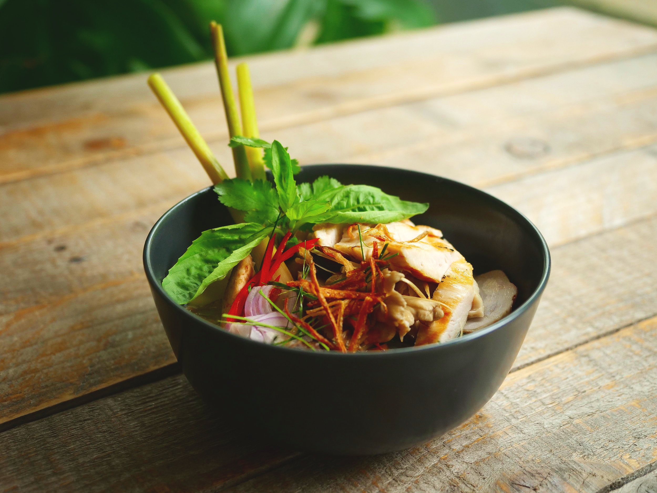Smoked chicken tom kha soup with wild mushrooms, sweet basil and lemongrass in a dark bowl on a wooden surface, recipe by Sebby Holmes