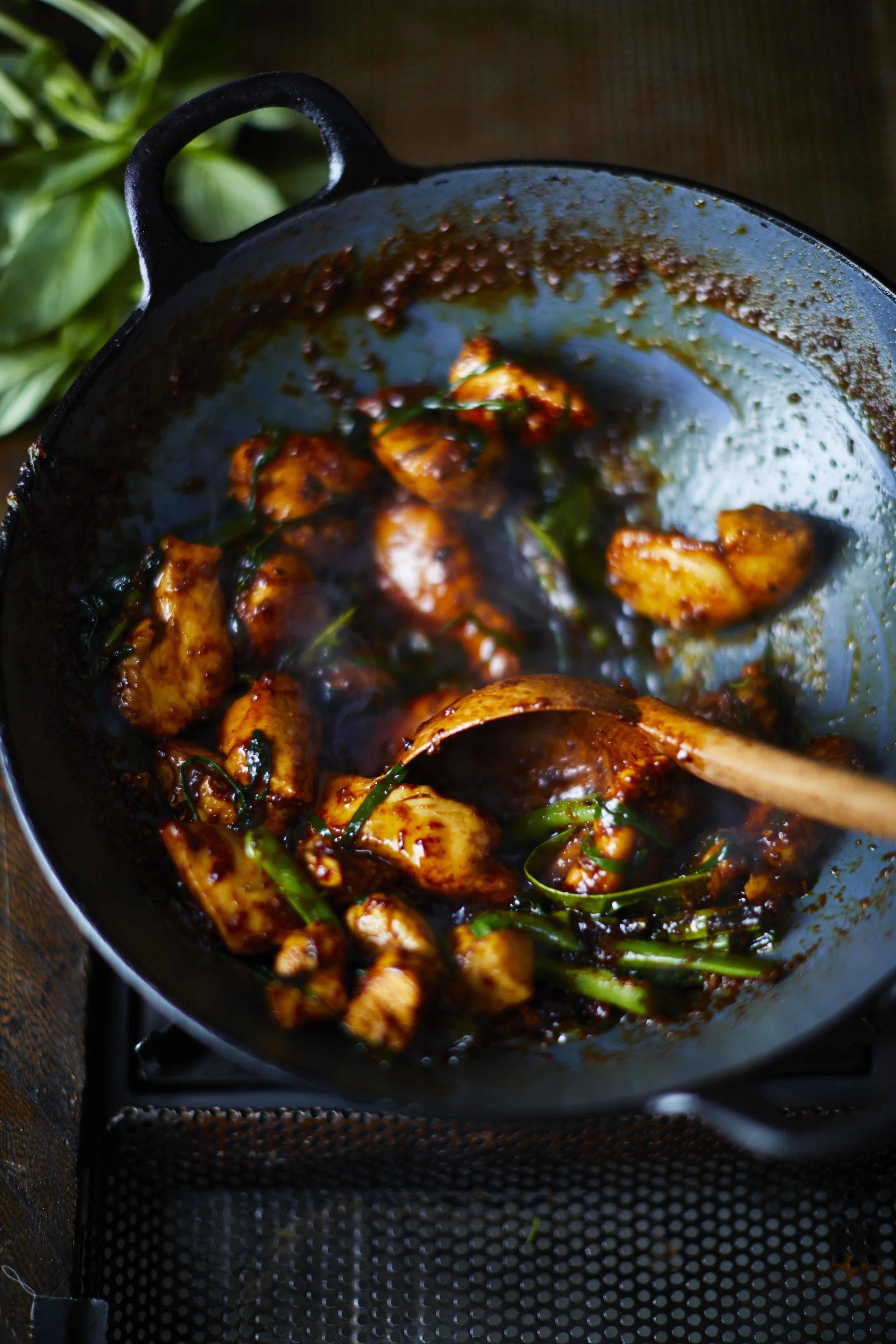 Stir-fried chicken pieces coated in dark nahm prik pao chilli jam with morning glory and green mango in a wok, recipe by Sebby Holmes